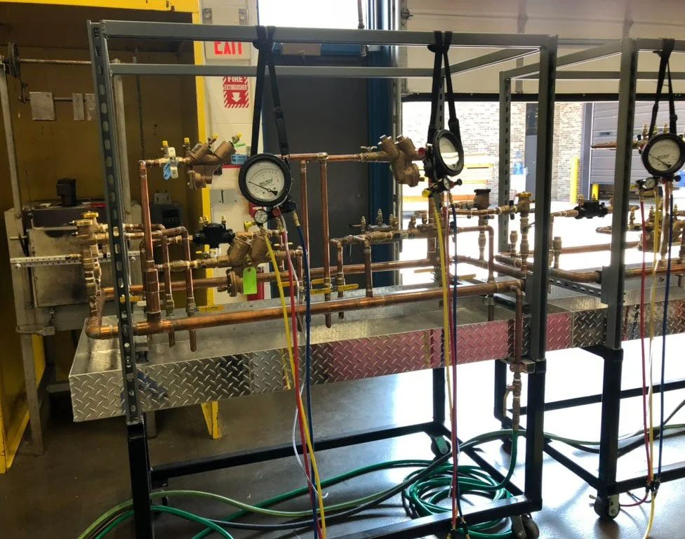 A mechanical training setup with copper pipes, gauges, and tubes on a mobile metal frame in a workshop or industrial setting.