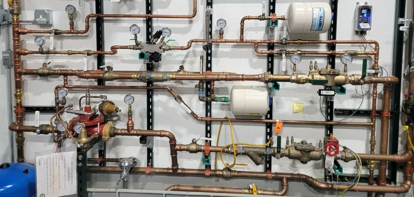 Complex plumbing system with numerous copper pipes, gauges, valves, and white tanks mounted on a wall.