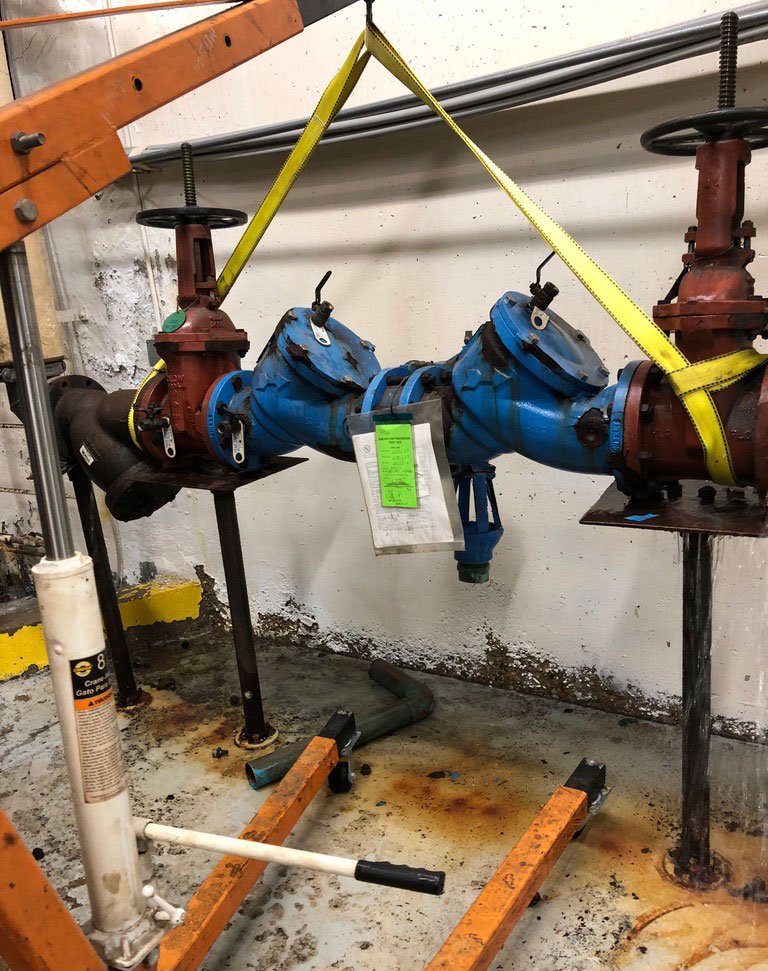 Large blue industrial valve or pipe fitting connected to red pipes, secured with yellow straps, in an industrial setting with rusted floor and tools.