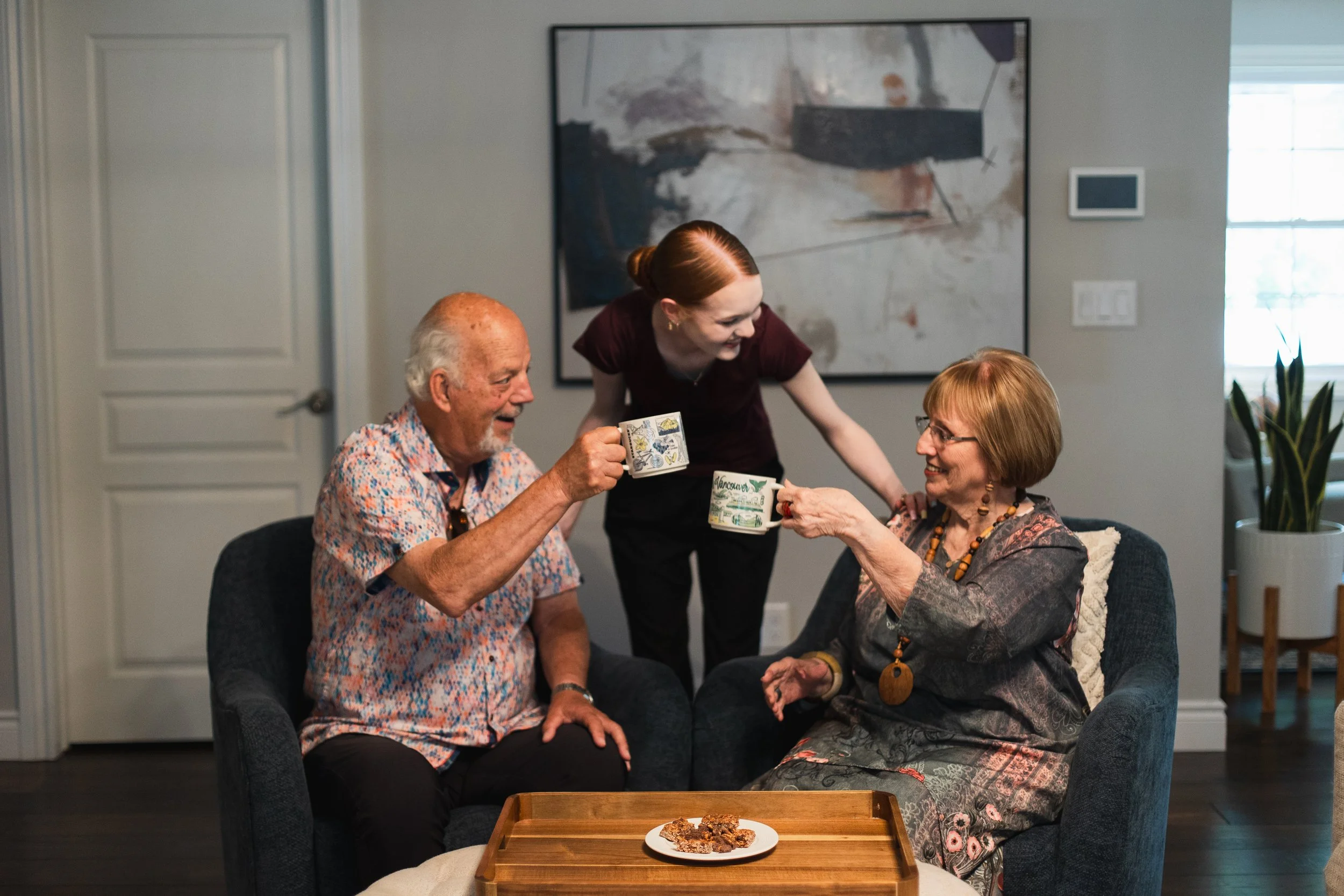 Better Days companion care for seniors in Kitchener-Waterloo. An elderly man and woman sitting on a couch sharing a mug of tea or coffee, smiling at each other, while a woman pours a drink for the woman. 