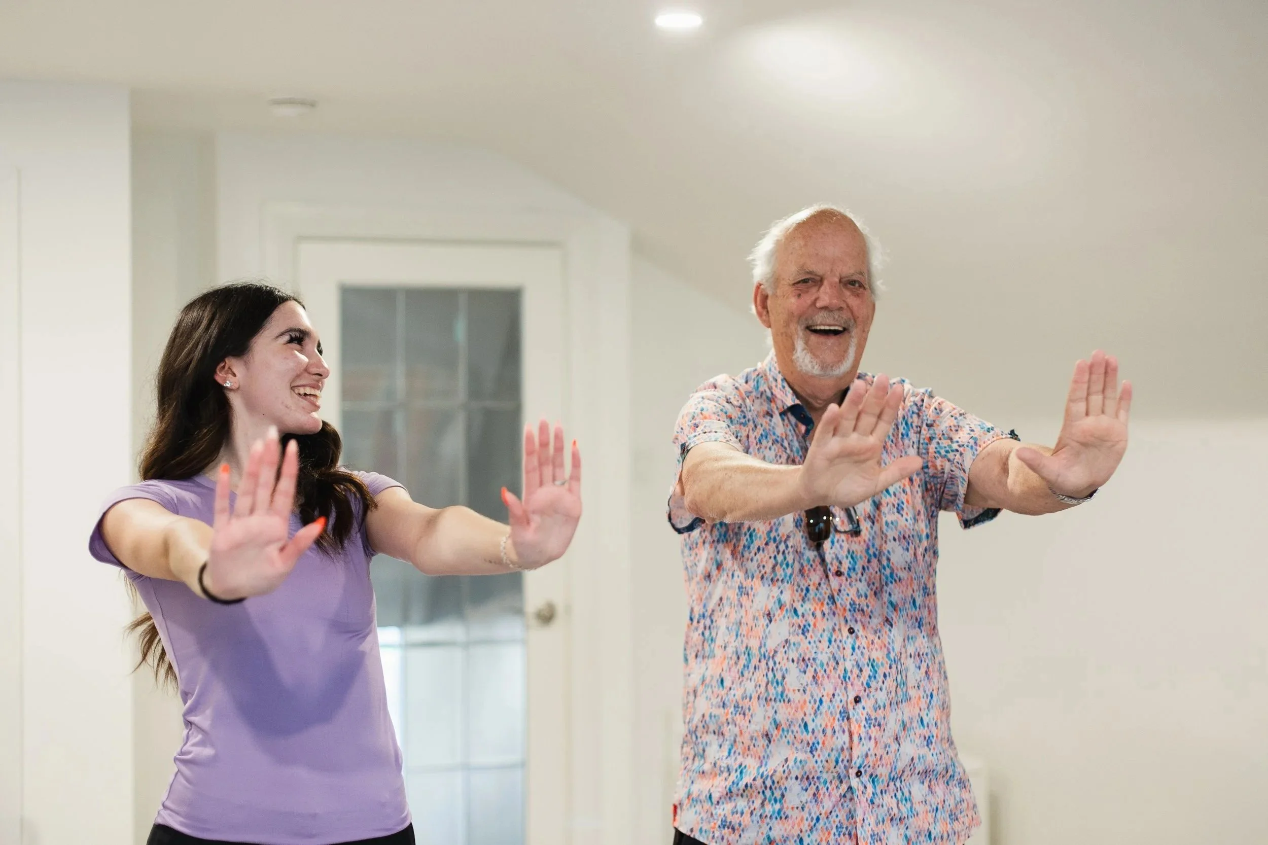 Better Days companion care for seniors inA young woman and an older man are engaging in a dance or exercise class, standing side by side with their arms extended and palms facing forward, smiling and enjoying the activity in a bright, indoor setting.