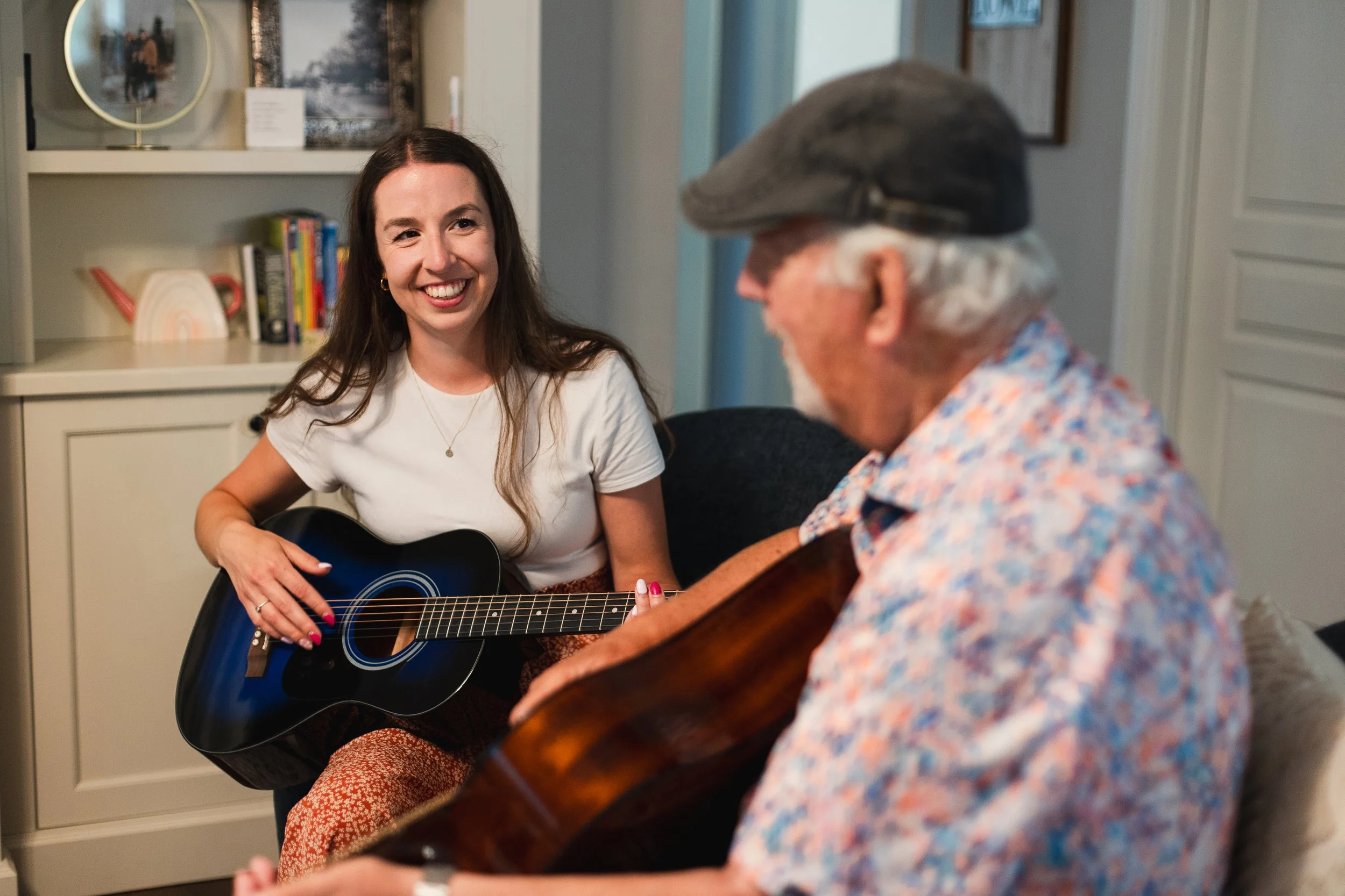 Better Days companion care for seniors in Kitchener-Waterloo. A young woman with long dark hair smiling while playing a black acoustic guitar, sitting across from an older man with white hair and a beard.