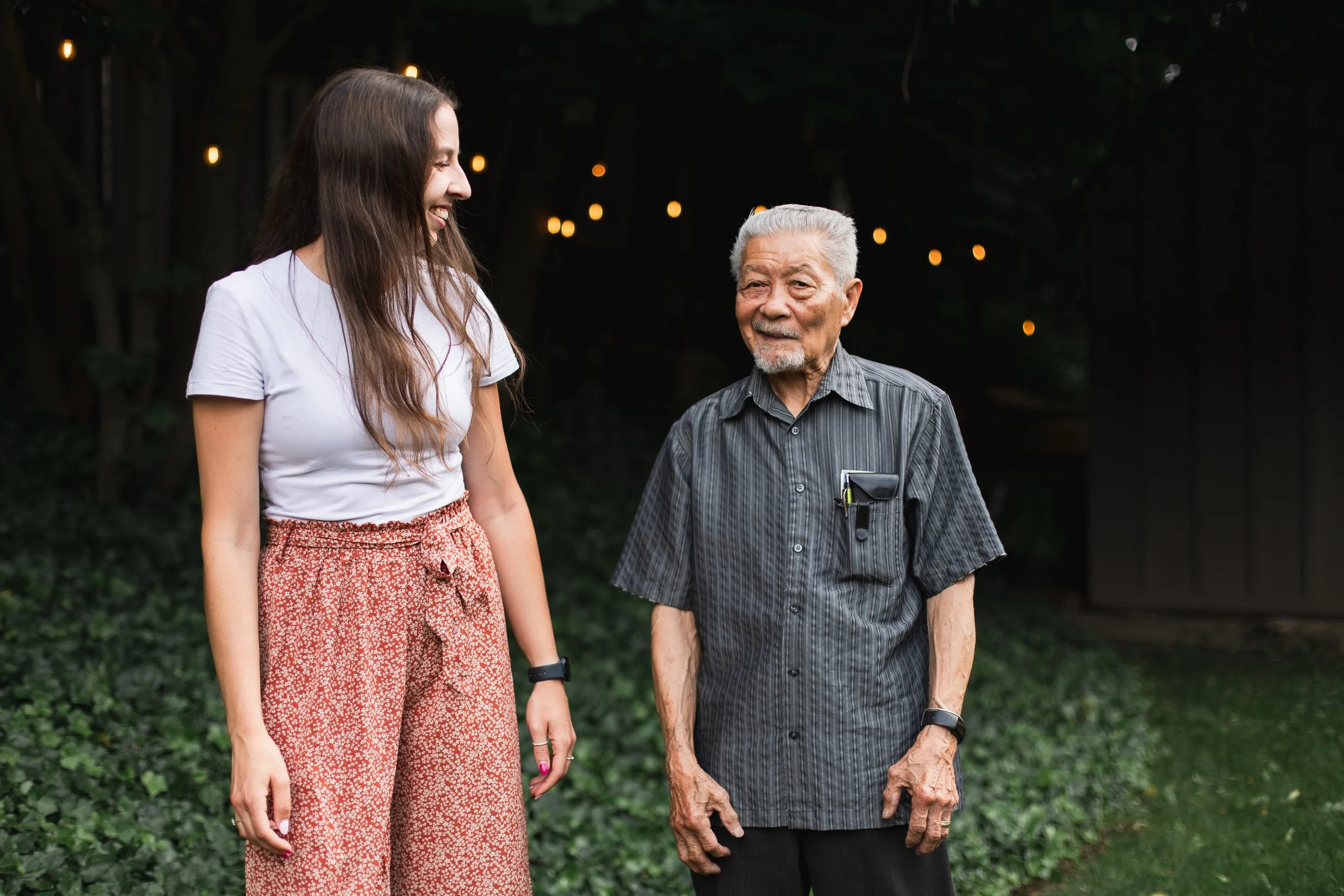 Better Days companion care for seniors in Kitchener-Waterloo. A young woman with long brown hair smiling and looking at an elderly man with white hair and a beard, who is wearing a striped black shirt. 
