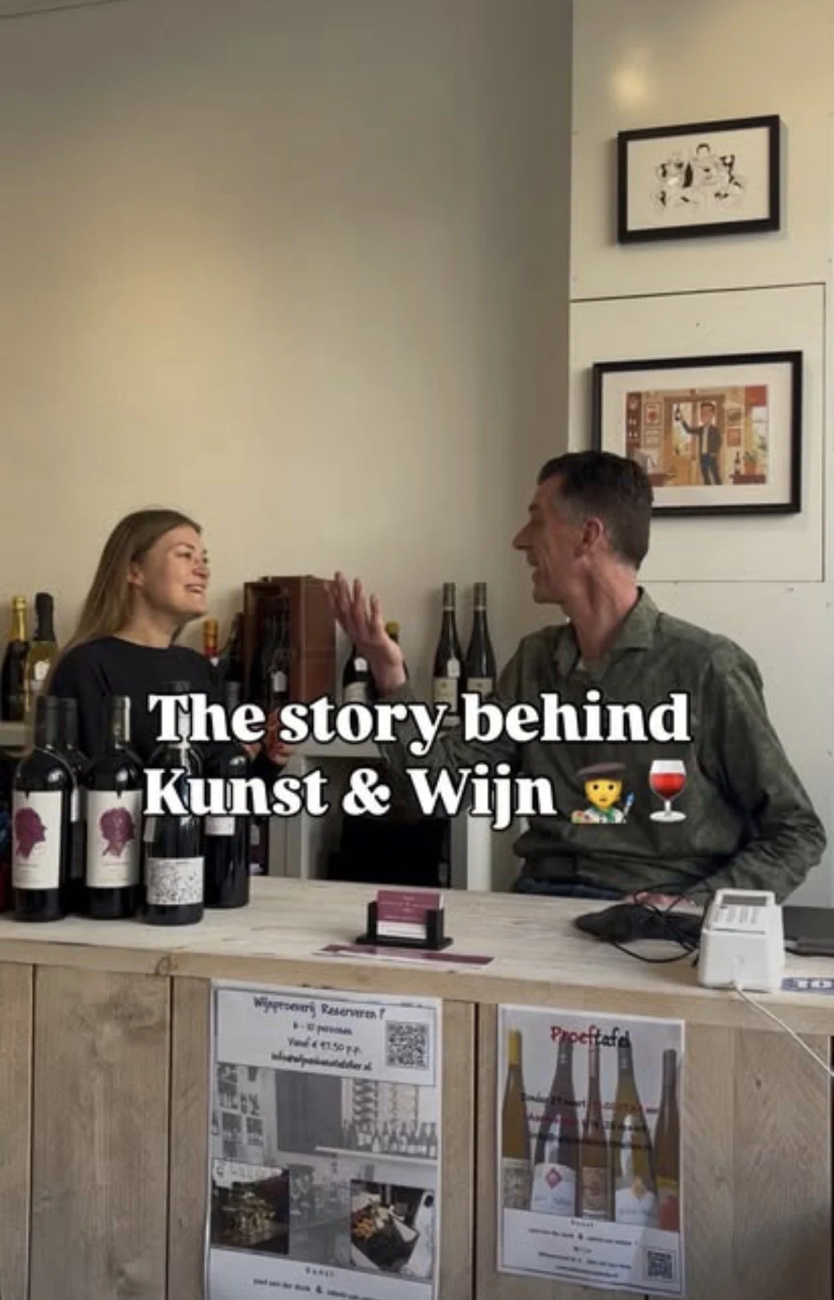 Two people, a woman and a man, talking and smiling at a wine tasting event, with bottles of wine on the table and framed pictures on the wall in the background. The text on the image reads 'The story behind Kunst & Wijn' with a wine glass emoji.