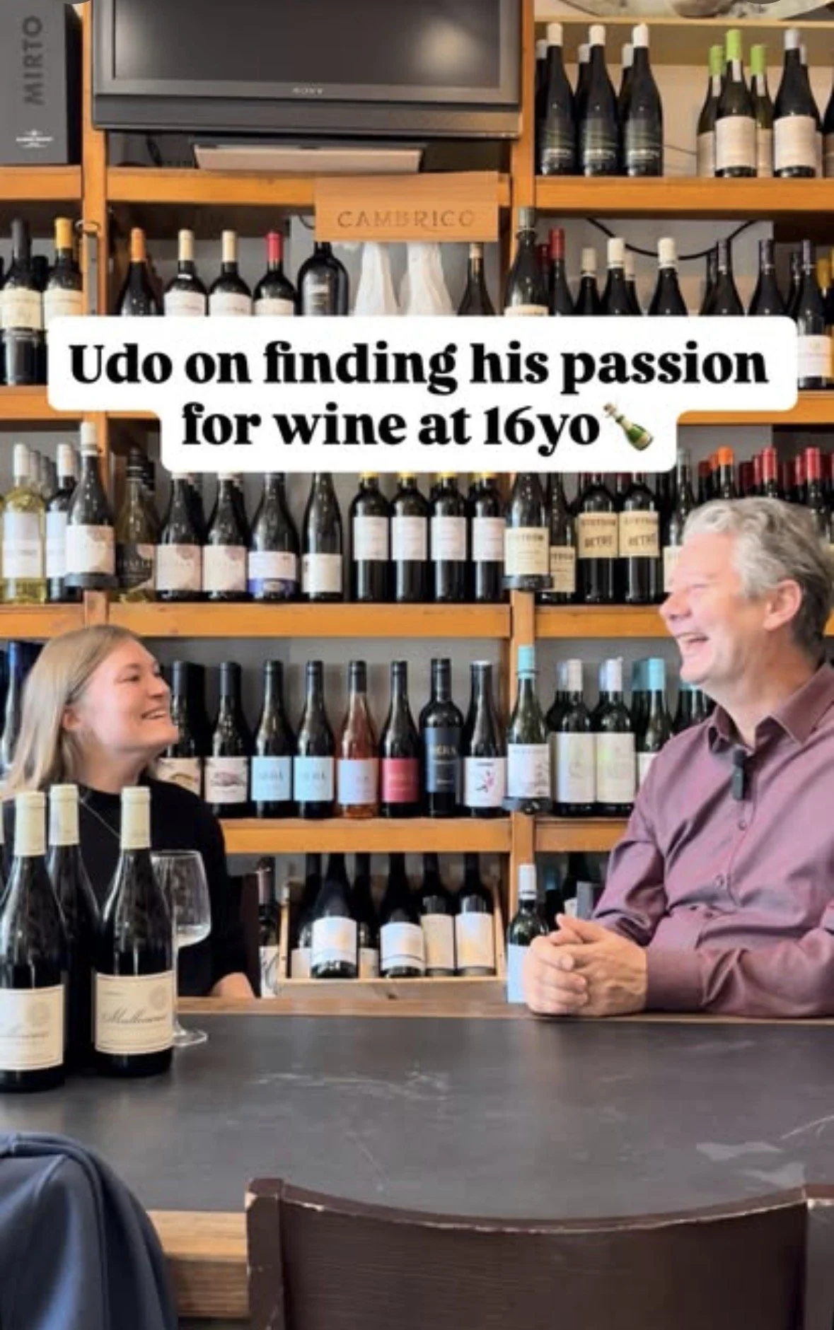 Two people, a woman and a man, are smiling and talking at a wine bar. Behind them, shelves are filled with bottles of wine. They are talking about Winematters.nl