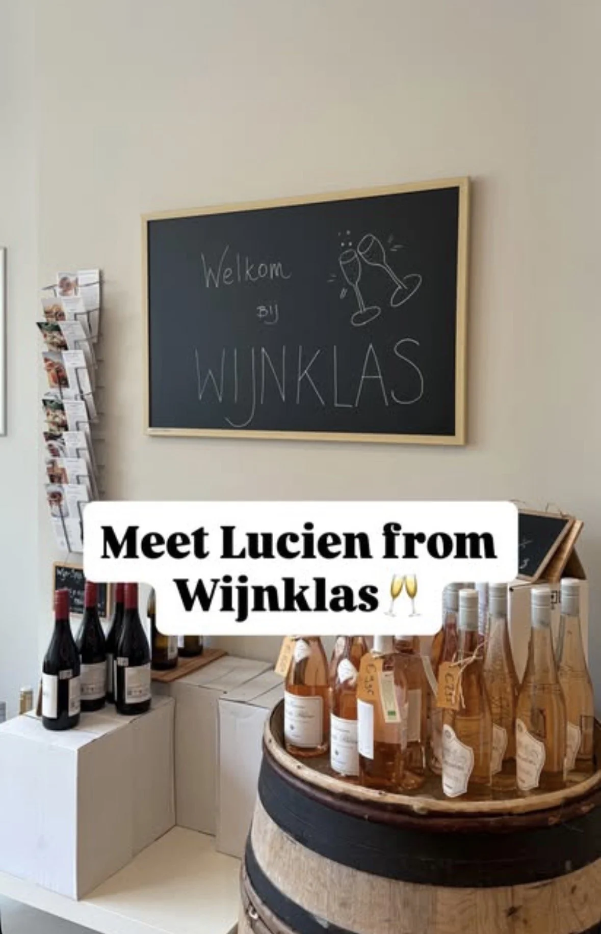 Interior of a wine store with a chalkboard sign saying "Welkom bij Wijnkas" and illustrations of clinking wine glasses. There are bottles of wine and rosé displayed on tables and shelves.