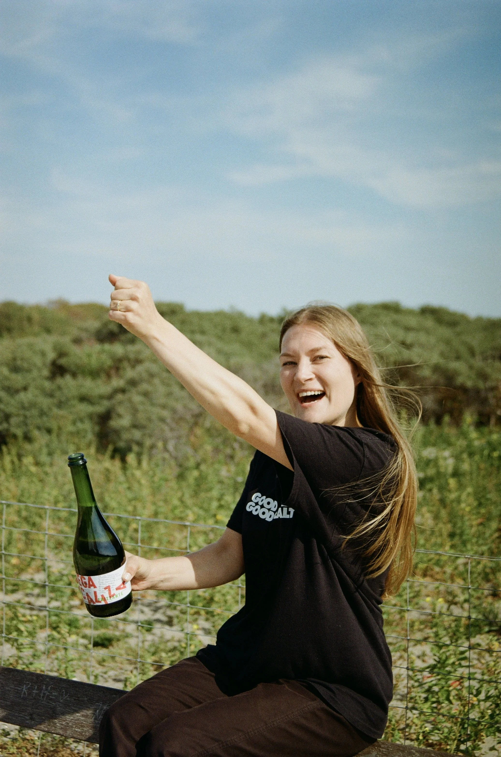 A woman sitting on a bench outdoors, holding a bottle of soda, smiling, and raising her arm in a celebratory gesture. She has long hair, is wearing a black t-shirt with white text, and brown pants. There are green bushes and a blue sky in the background.