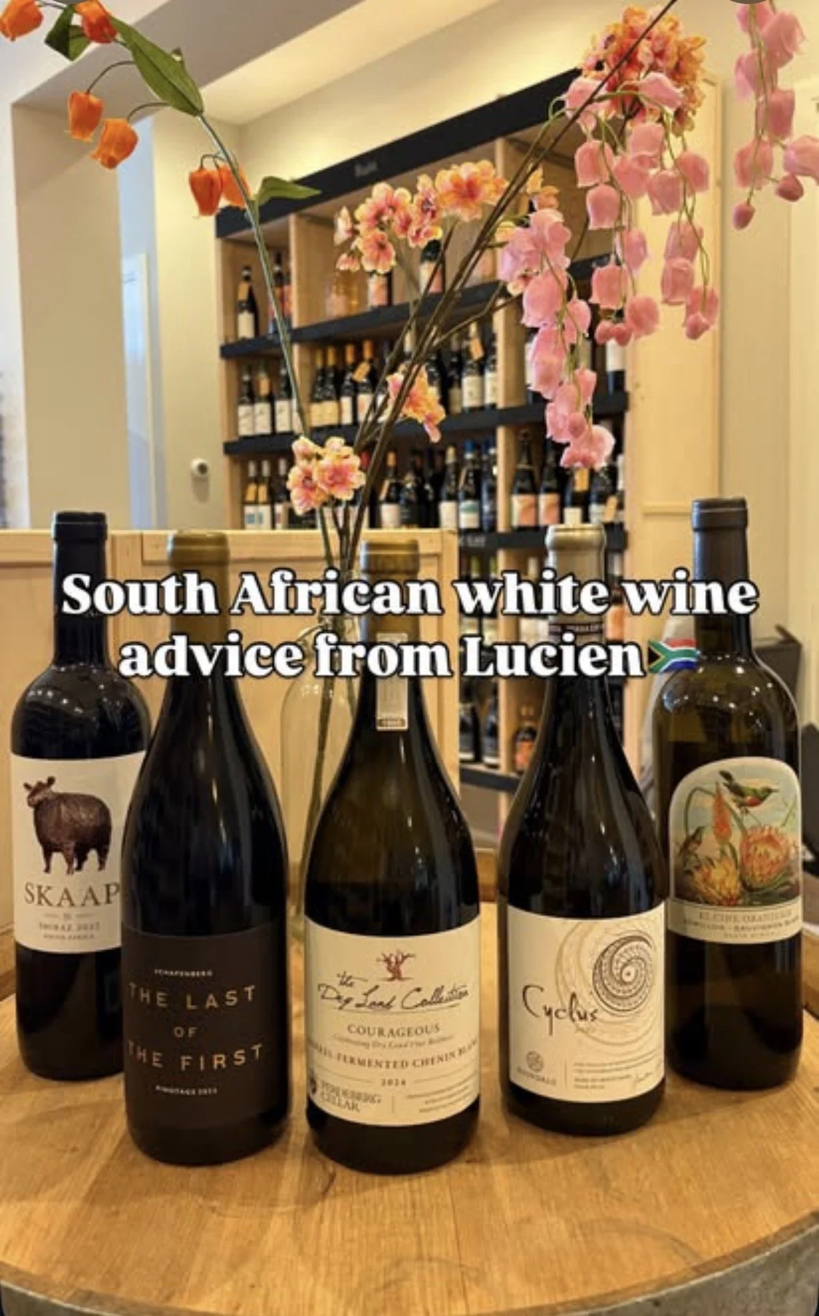 Four bottles of wine on a wooden table with pink flowers in a vase behind them, and a wine shelf in the background. Text overlay reads 'South African white wine advice from Lucien'. Wijnklas advice for wine.