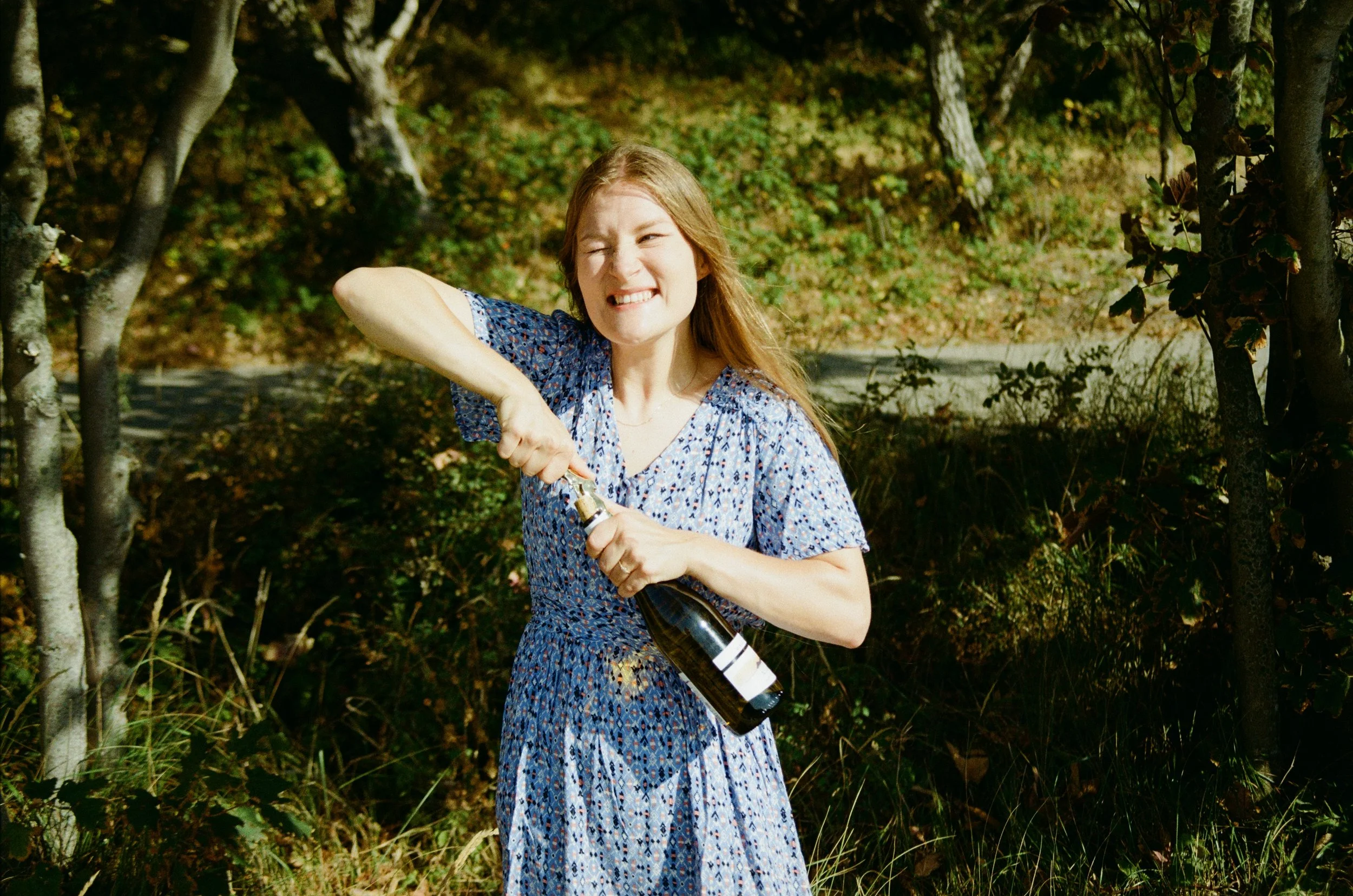 A young woman with long red hair smiling and holding a bottle of beer outdoors in a wooded area on a sunny day.