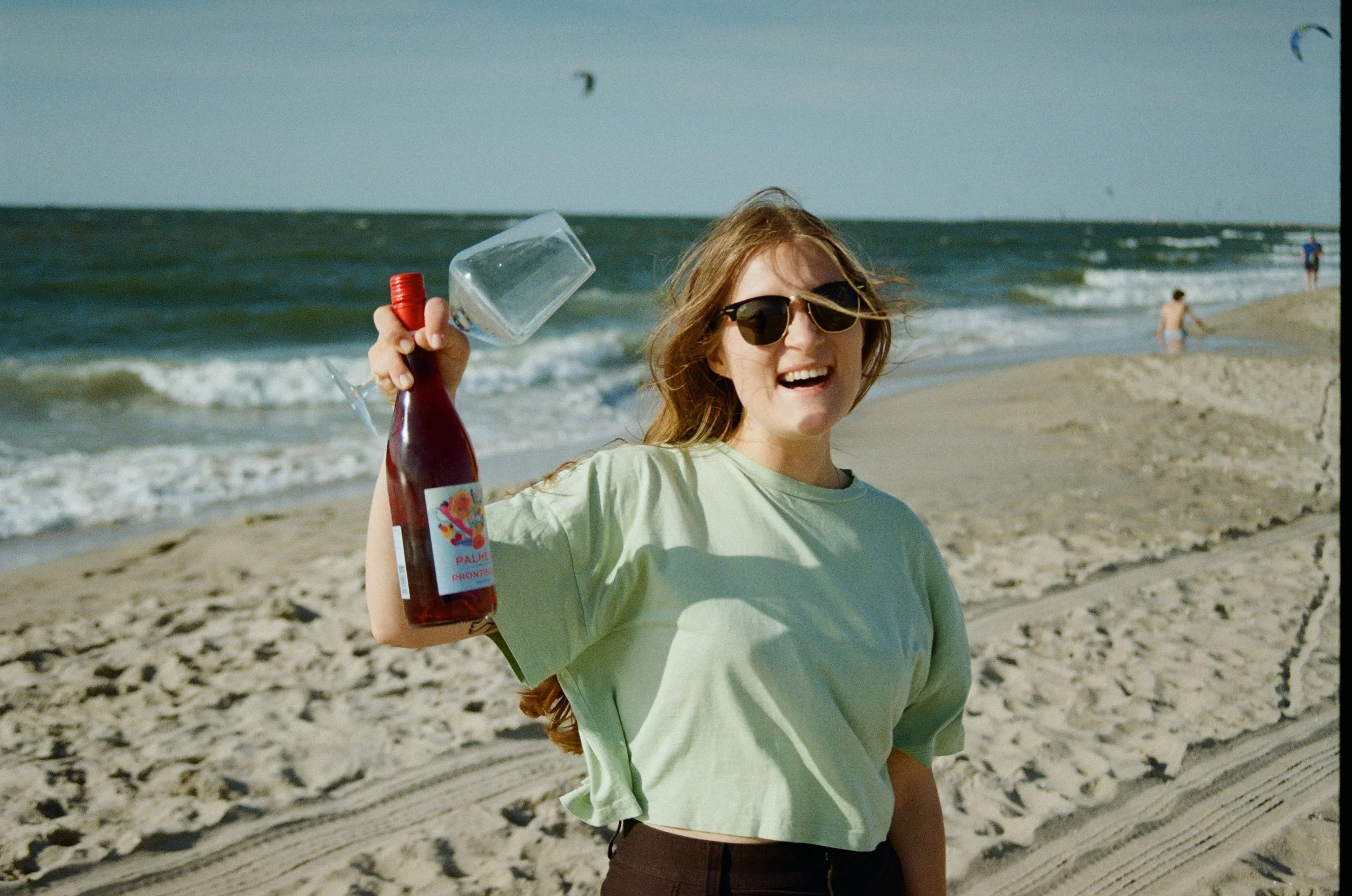 A smiling woman with sunglasses holding a bottle and an empty wine glass on a beach with waves.