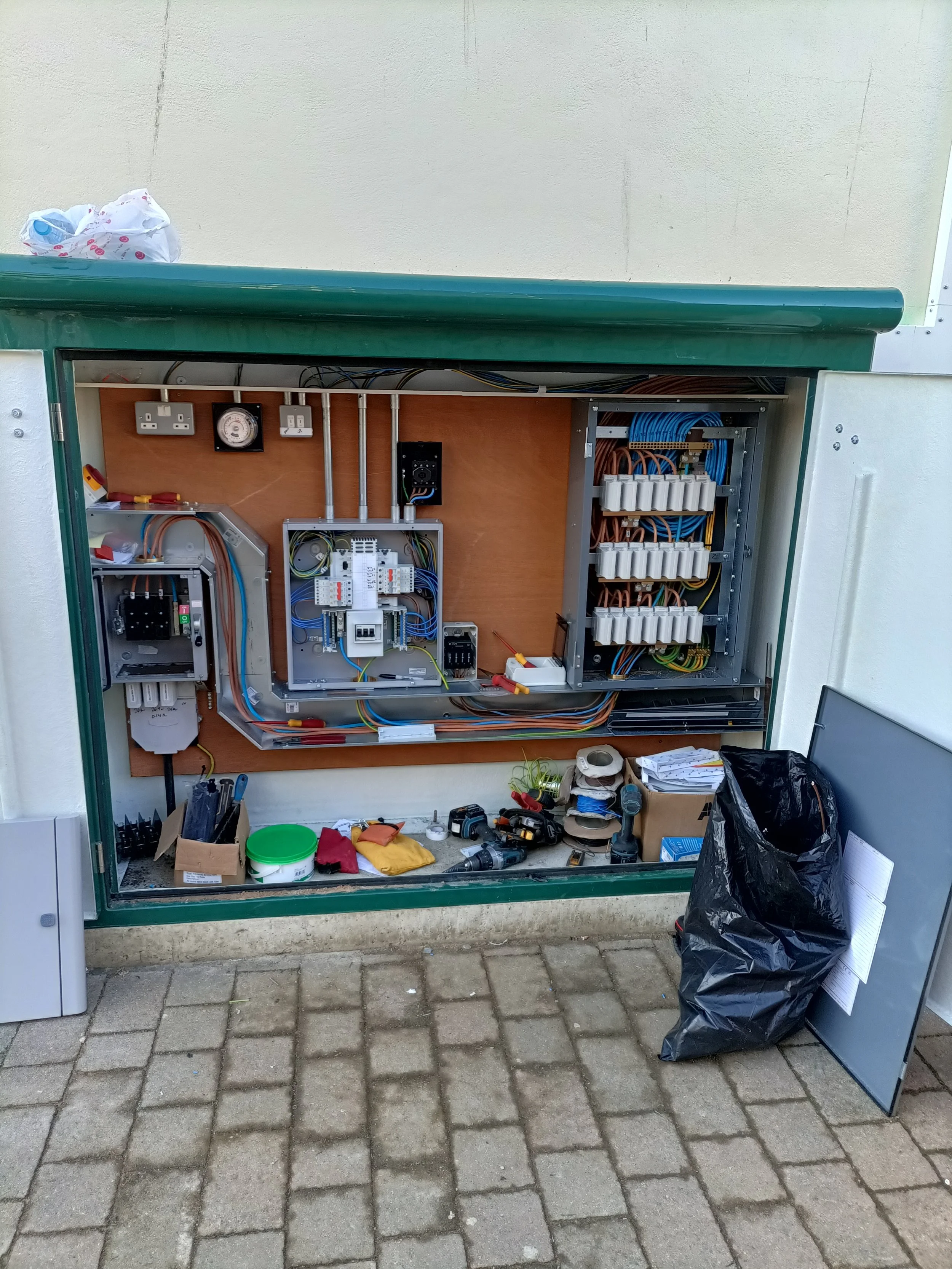 Open electrical panel with wiring and tools on a workbench outside, surrounded by a black trash bag, papers, and small objects on a brick-paved ground.