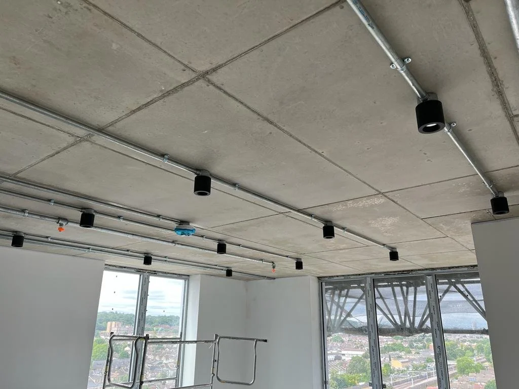Ceiling with exposed pipes and black light fixtures in a room with large windows showing a cityscape outside.