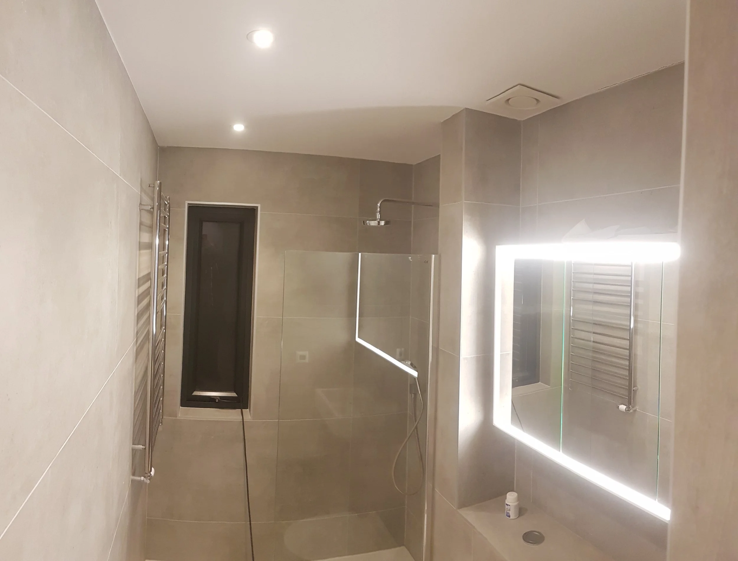 A modern bathroom with beige tiles, a wall-mounted mirror with LED lighting, a showerhead, a towel radiator, and a bottle of lotion on the sink.