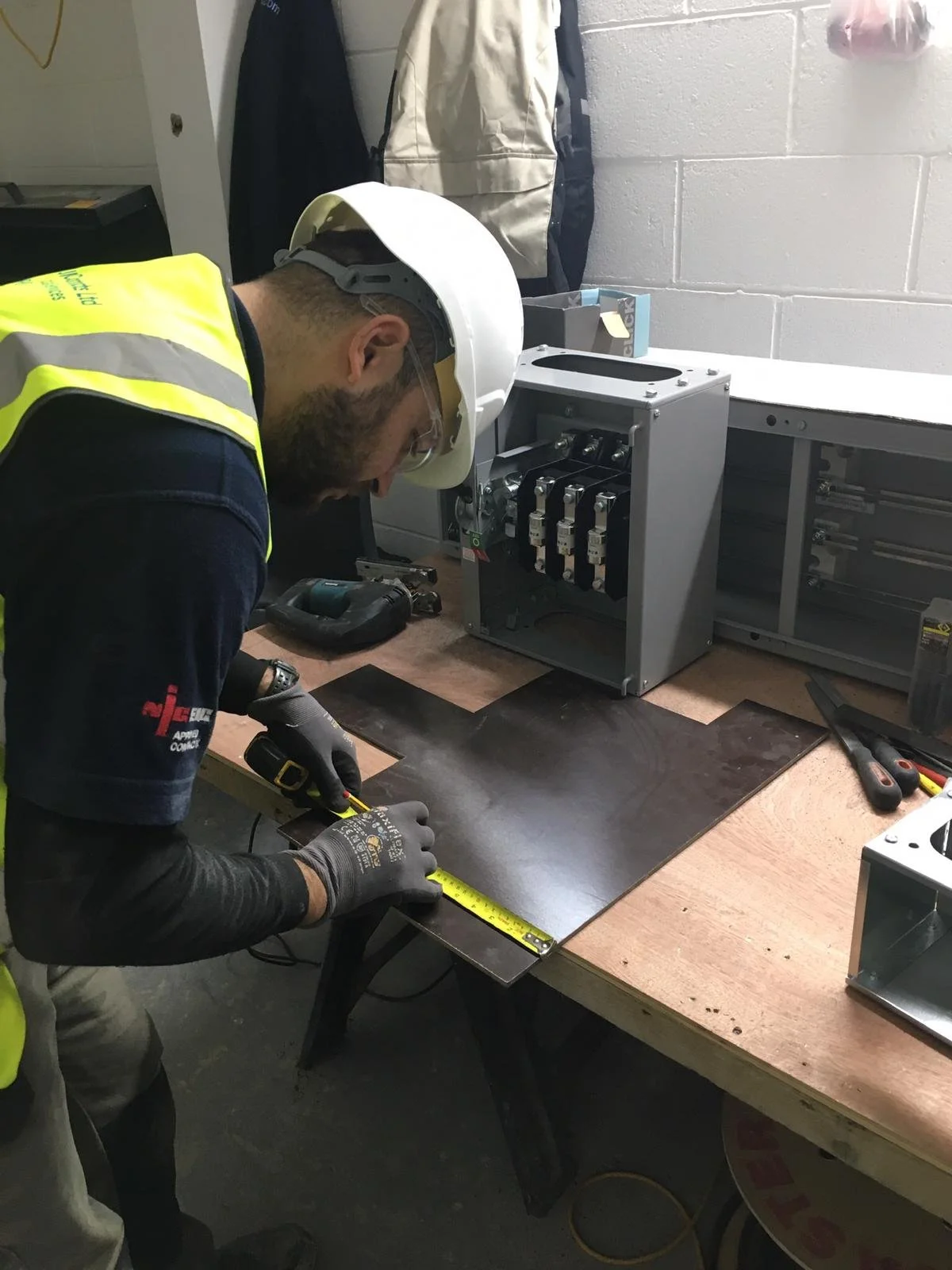 A worker wearing safety glasses, a hard hat, and gloves is measuring a wooden board with a tape measure on a workbench. The workspace has electronic components, tools, and equipment.