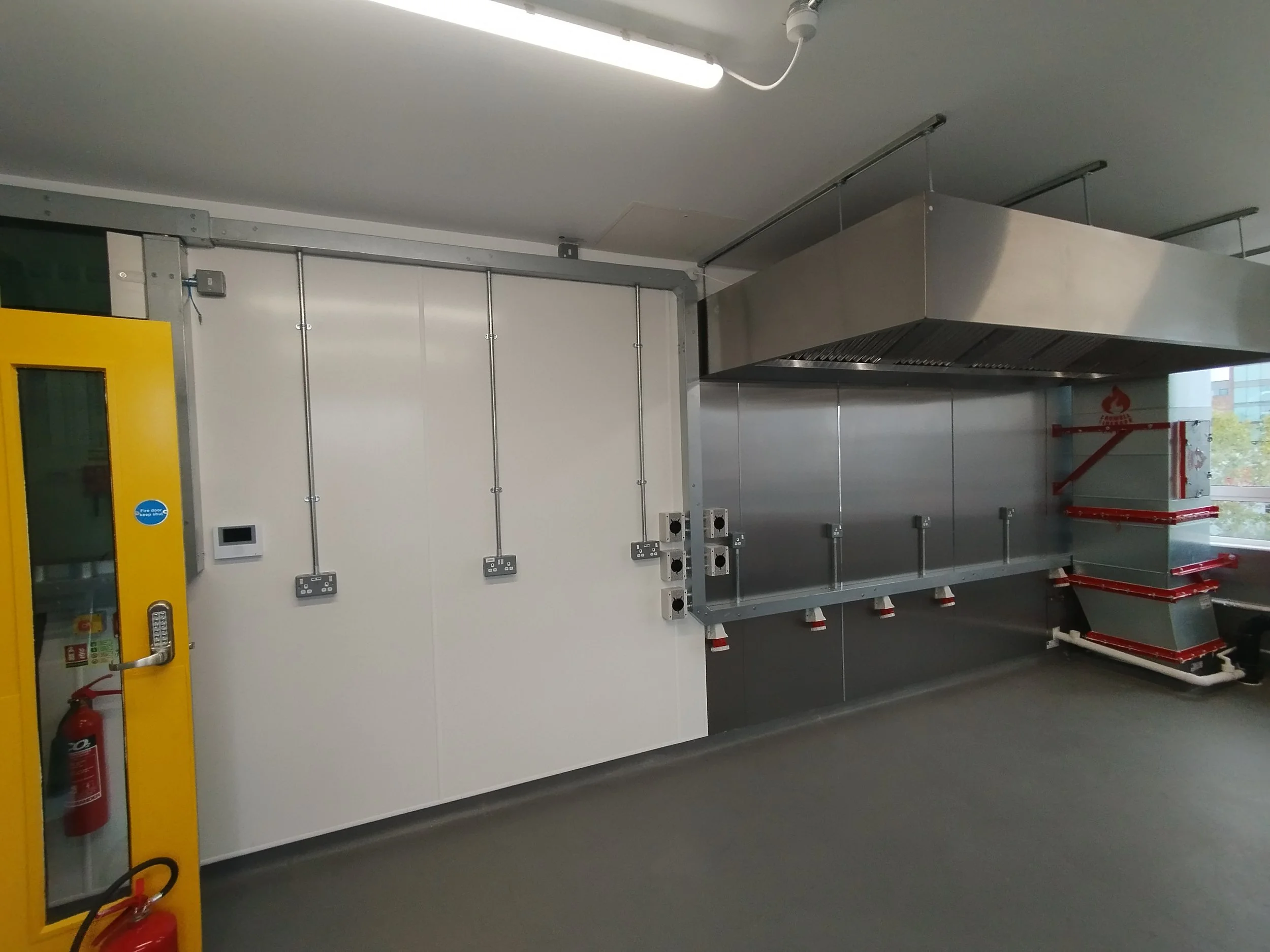 An interior of a commercial or industrial kitchen area shows a large stainless steel exhaust hood above a cooking or preparation area. There are electrical outlets and switches mounted on the wall, a yellow fire door with a window, and a fire extinguisher on the wall. The ceiling has fluorescent lighting.