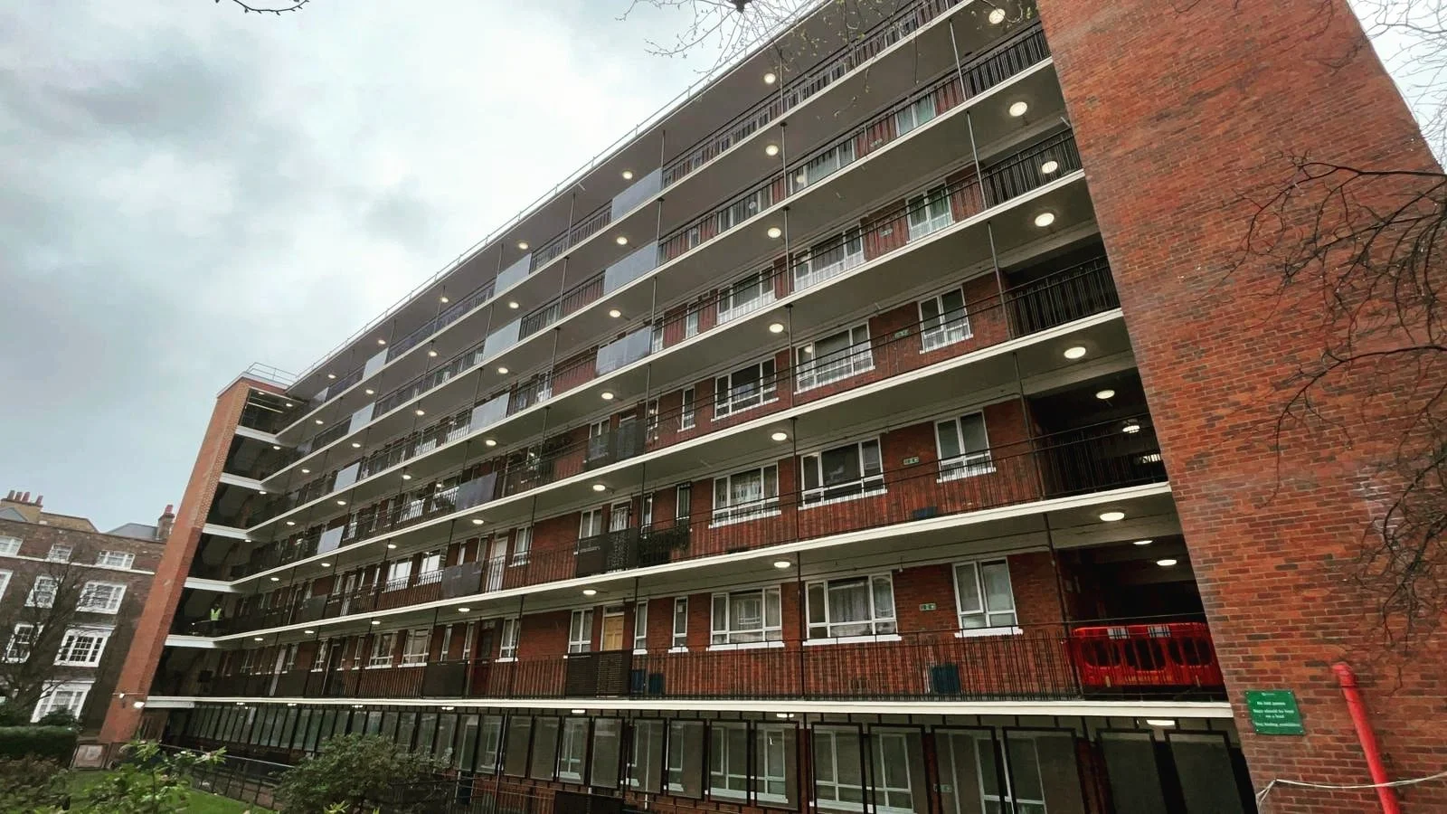 Multi-story residential building with brick facade, several balconies, some with outdoor furniture, and illuminated ceiling lights. Overcast sky and leafless tree branches in the foreground.