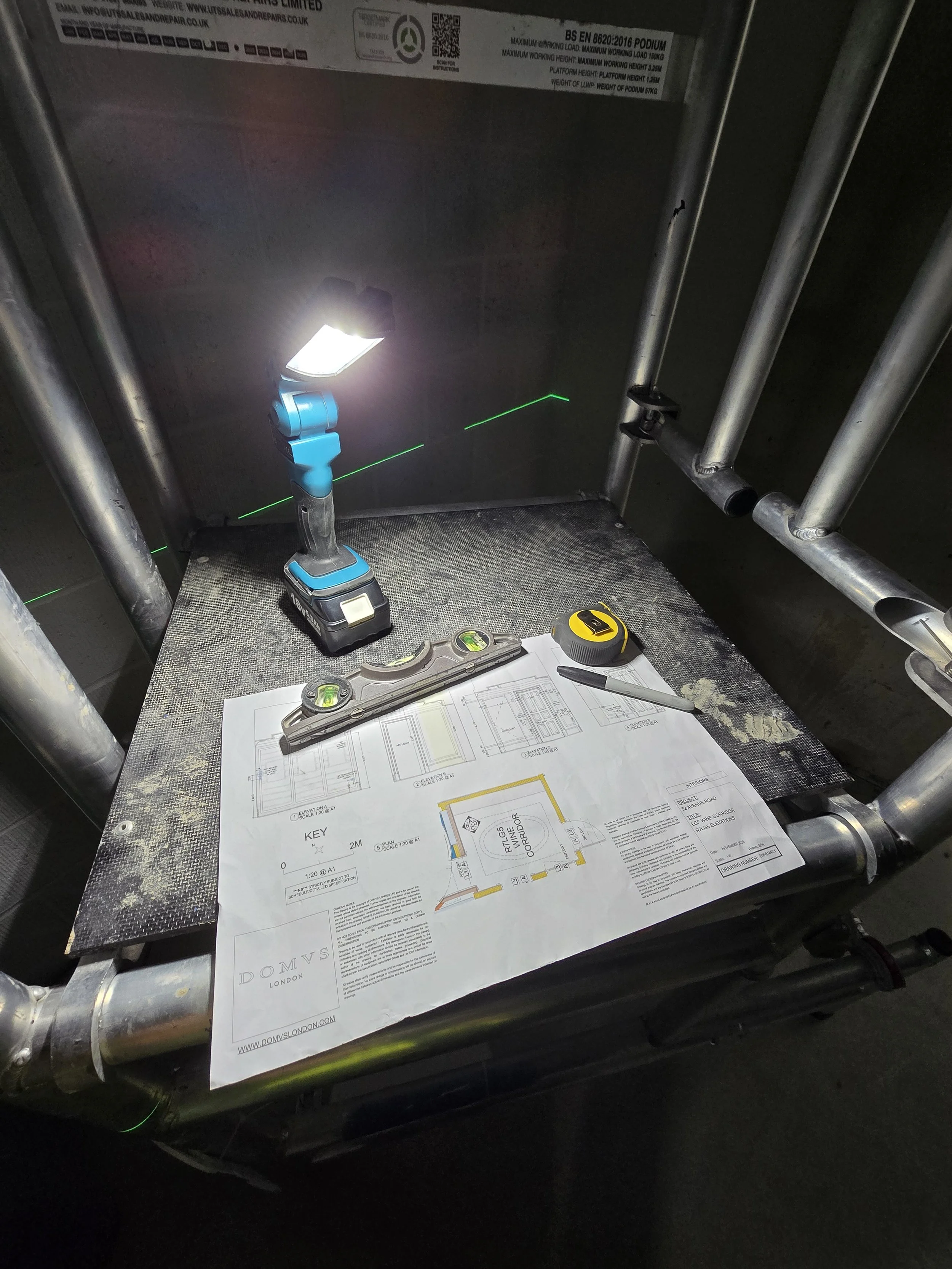 A work station with blue cordless drill, a yellow measuring tape, a spirit level, technical drawings, and writing tools on a surface made of textured metallic material. The background shows piping and a metal panel with technical labels.