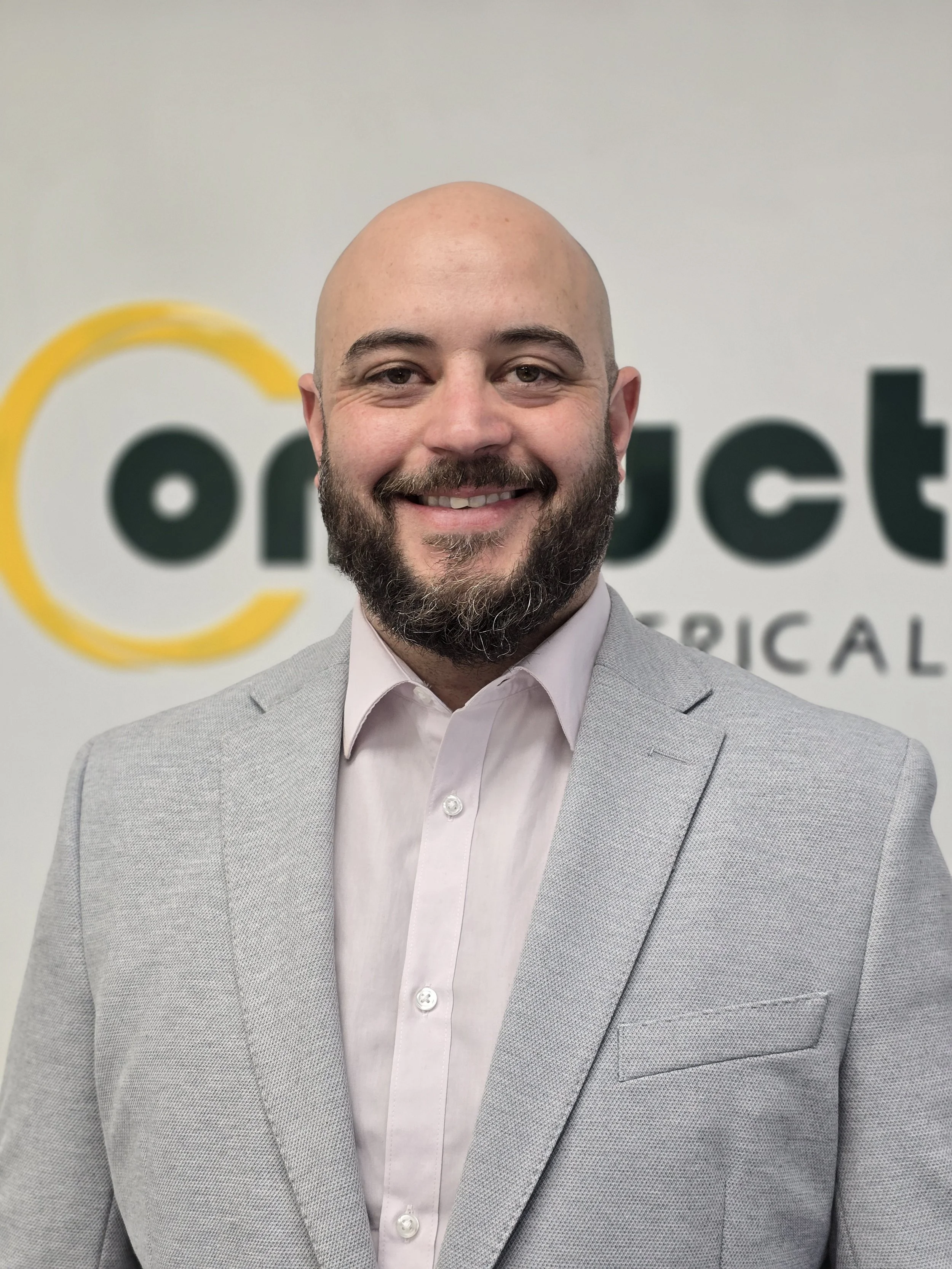 A smiling man with a beard and bald head, wearing a light gray suit and a pink dress shirt, standing in front of a wall with a partial logo.