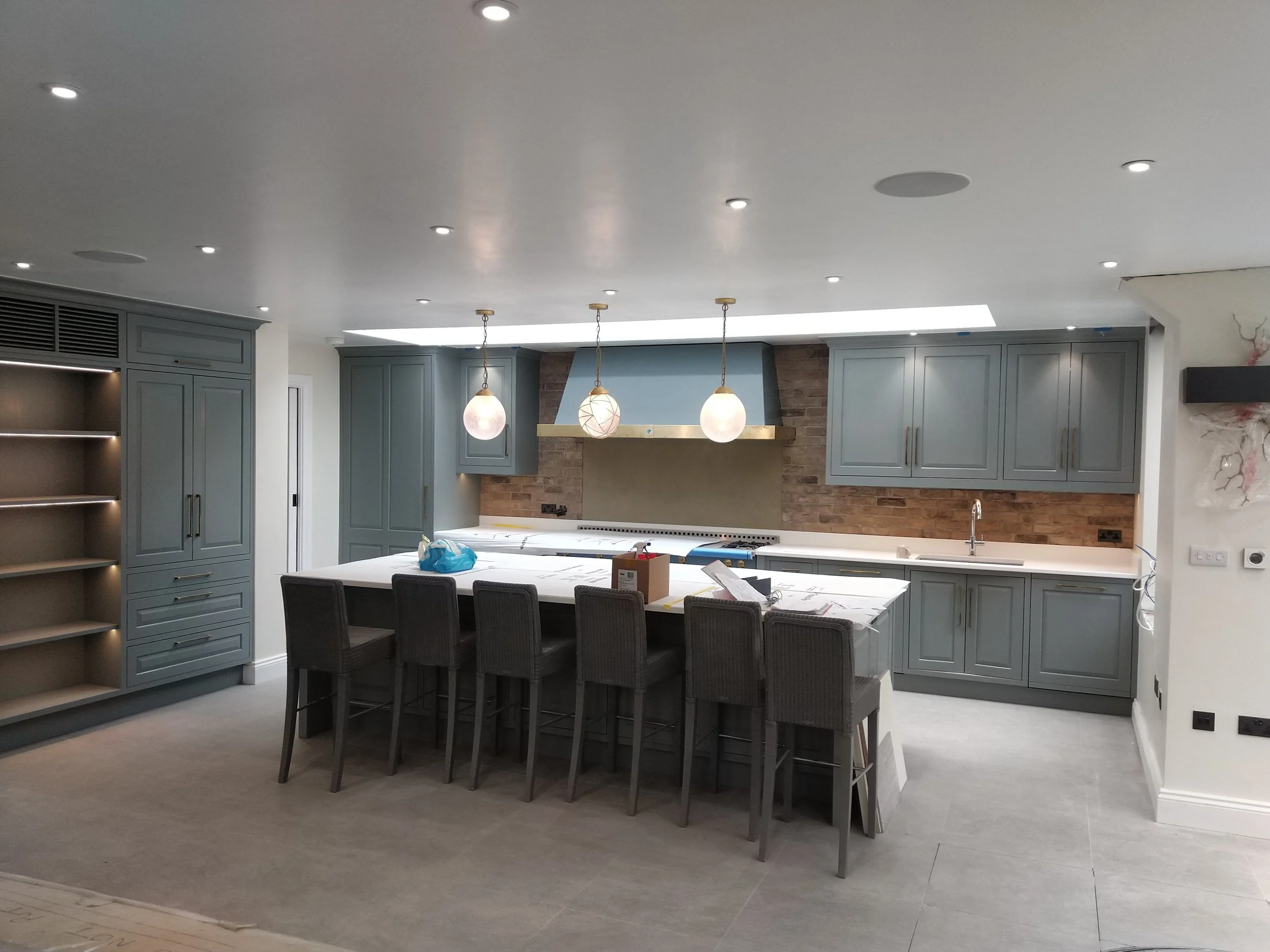 A kitchen with blue cabinets, a white island with chairs, pendant lights, a brick wall behind the stove, and a ceiling with recessed lighting.
