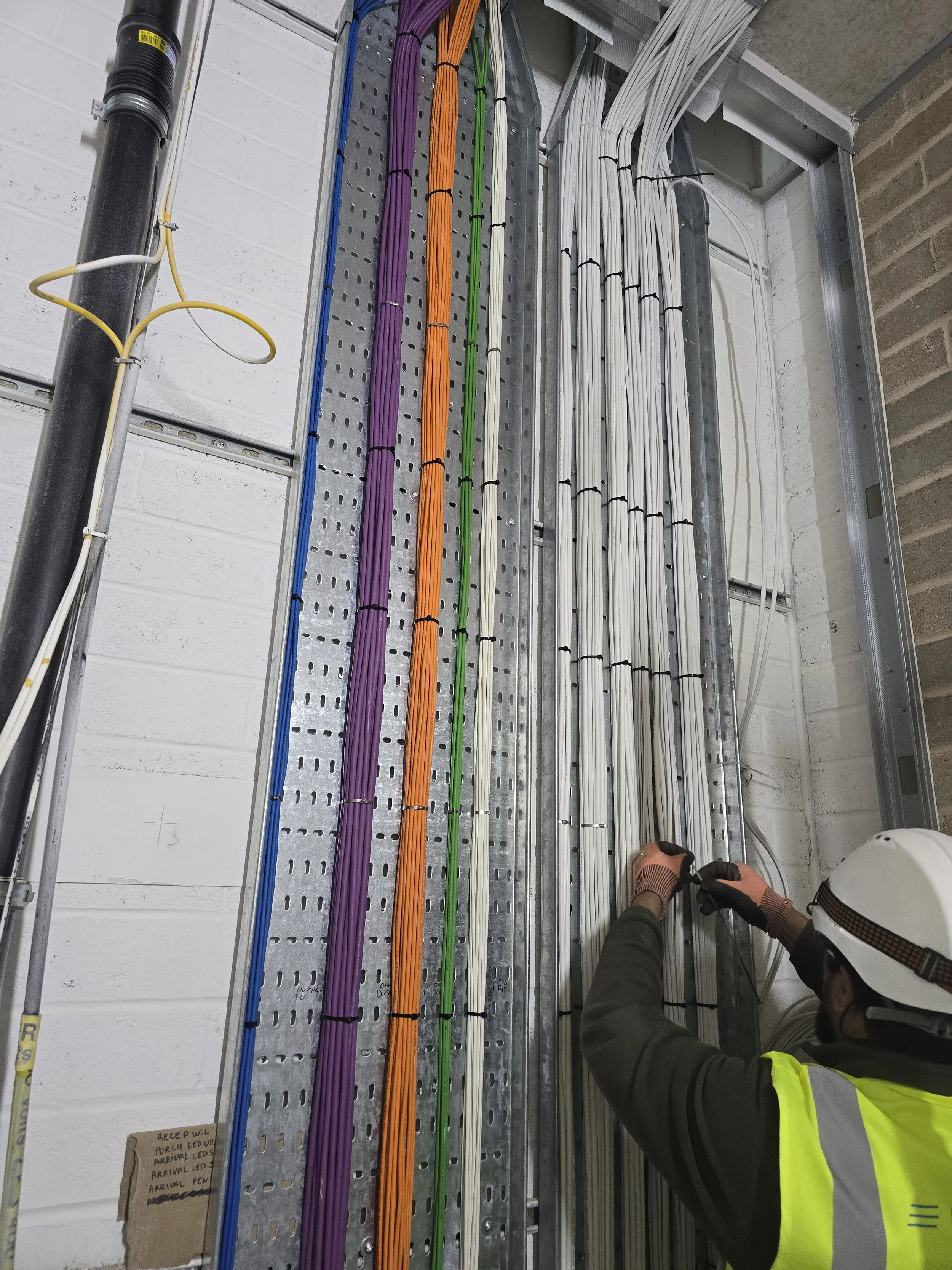 A construction worker wearing a hard hat, safety glasses, and a reflective vest is working on wiring inside a metal electrical panel with various colored cables organized and secured with black ties.