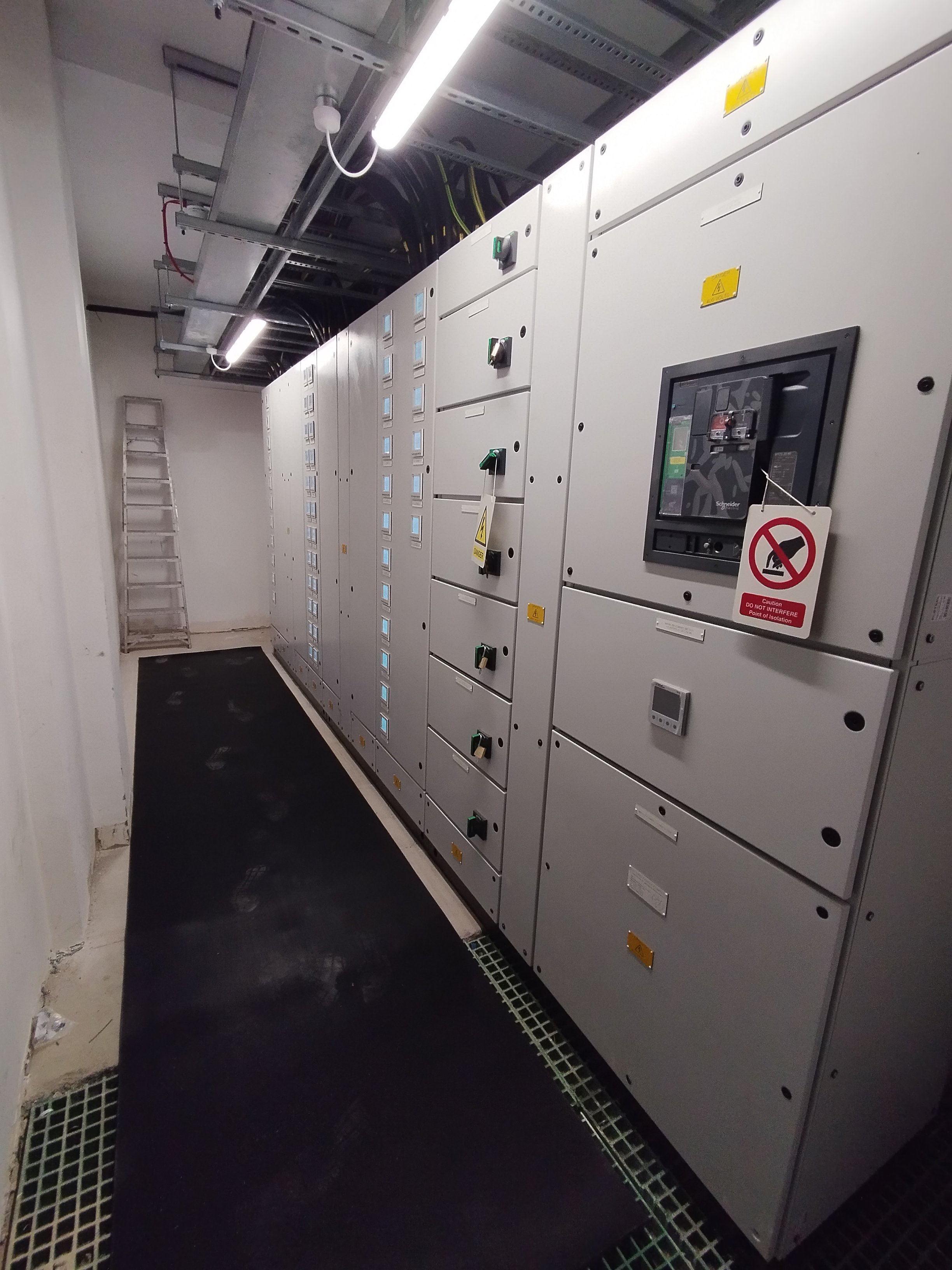 Electrical control panels with digital displays and switches in a room with a ladder against the back wall and a metallic cable tray above.