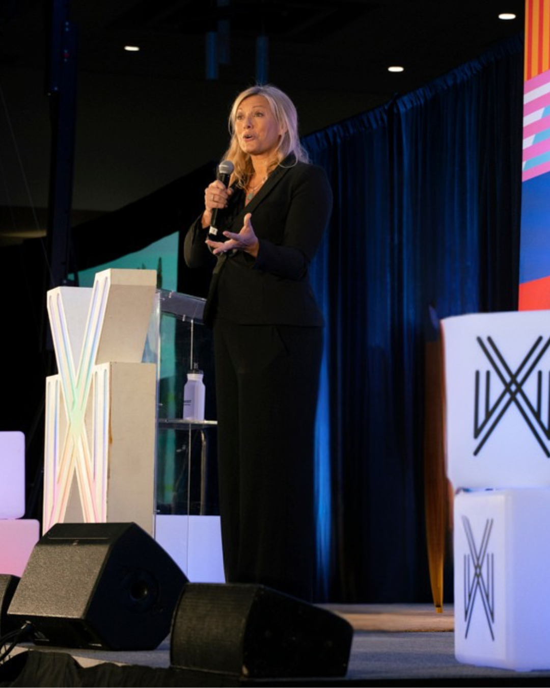 A woman in a black suit speaking into a microphone on stage at a conference or event, with a dark background, blue curtains, and colorful stage decorations.