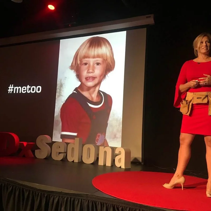 A woman in a red dress and beige heels standing on a stage with large letters spelling 'Sedona' and a large screen behind her displaying a child's portrait. The child has blonde hair and is wearing a red and black shirt. The stage has a black curtain
