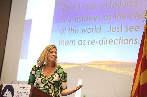 A woman standing at a podium with a microphone, giving a presentation. Behind her, a large screen displays a motivational message about seeing failures as directions. There is a flag to her right.