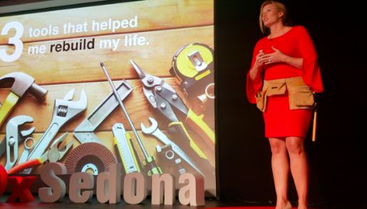 A woman in a red dress stands on stage next to a large screen displaying a slide about tools that helped rebuild her life, showing various tools like a wrench, hammer, screwdriver, and tape measure.