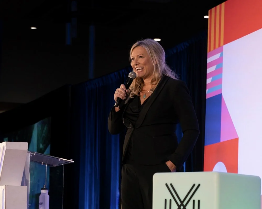 A woman with blonde hair, wearing a black blazer, speaking into a microphone on stage at a conference or event, with a large colorful screen behind her.