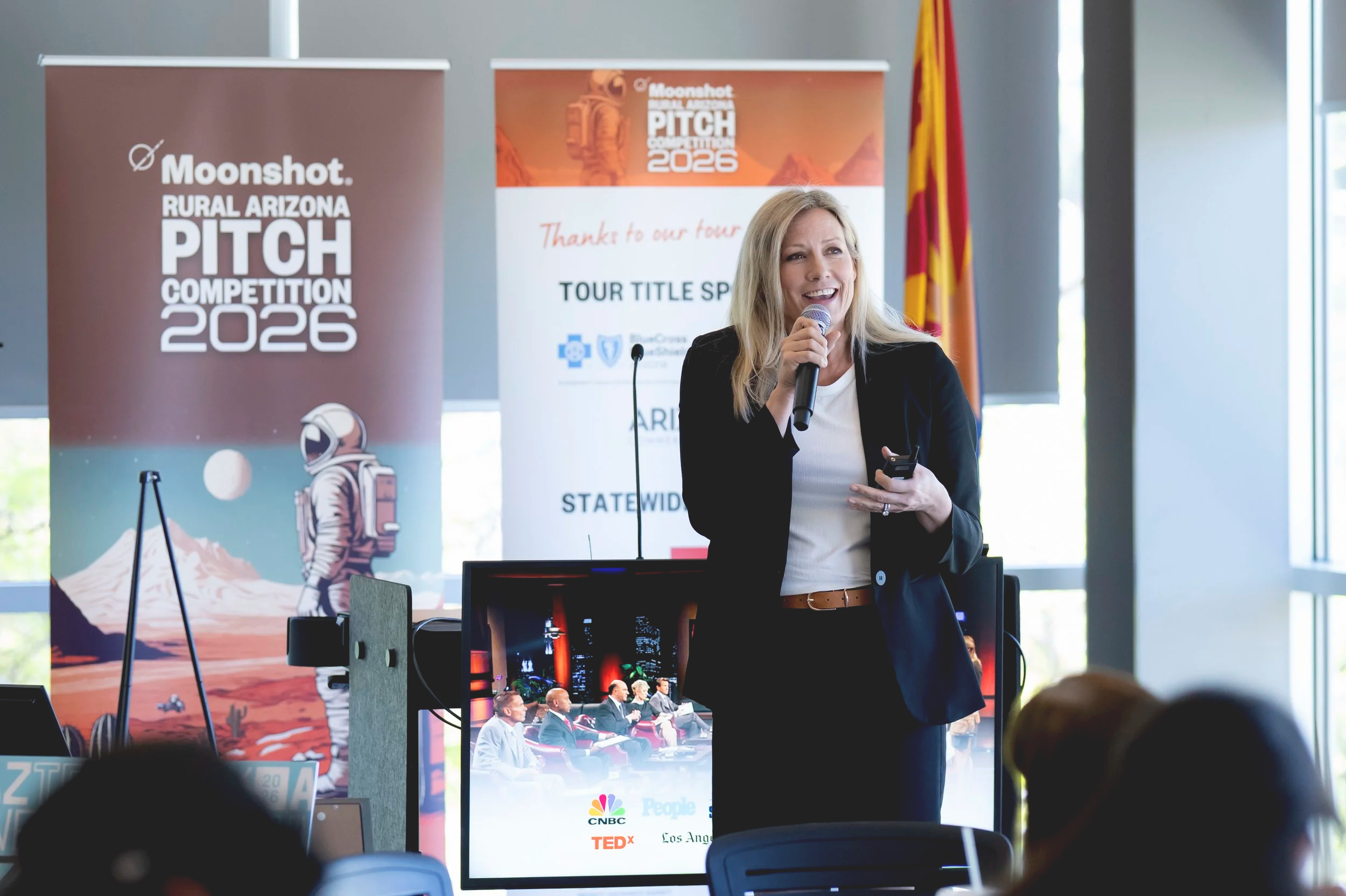 A blonde woman speaking into a microphone at a rural Arizona pitch competition event, with banners displaying the event's logo and name behind her, and an American flag to the right.