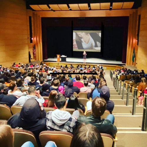 Audience attending a presentation or performance in a large auditorium, with a person on stage and a large screen behind them.