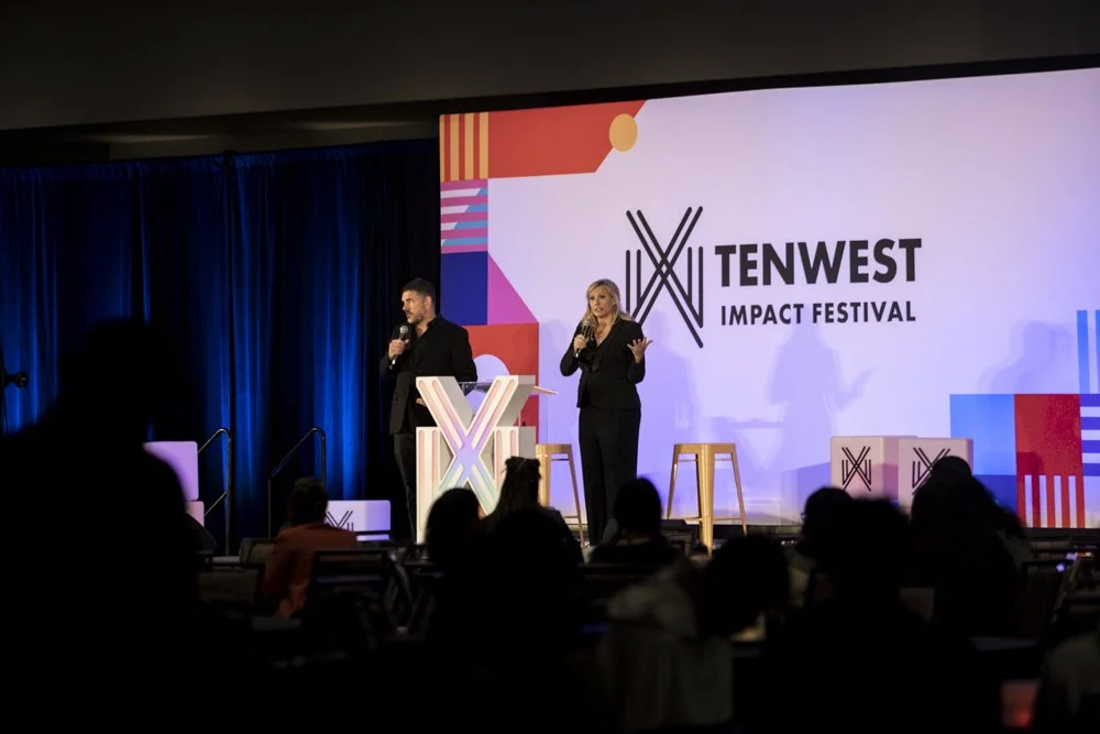 Two presenters on stage at the TenWest Impact Festival, speaking to an audience; the stage has a large screen with the festival's logo and colorful graphics.