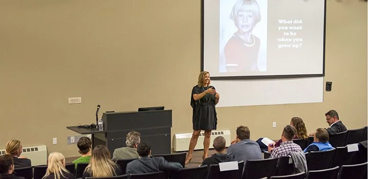 A woman giving a presentation in a classroom or conference room with an audience, a large screen displaying a childhood photo of her and the question 'What did you want to be when you grew up?'