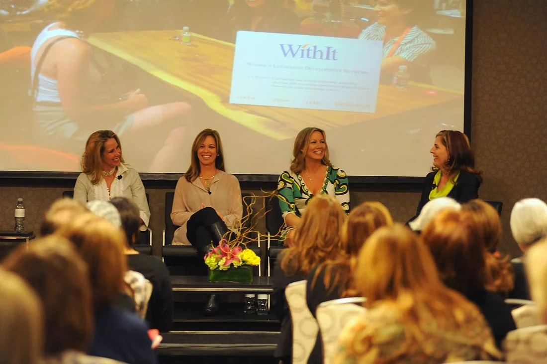 Four women sit on a panel during a conference, with a large screen behind them displaying the word 'WithIt' and other text. The audience women are visible in the foreground.