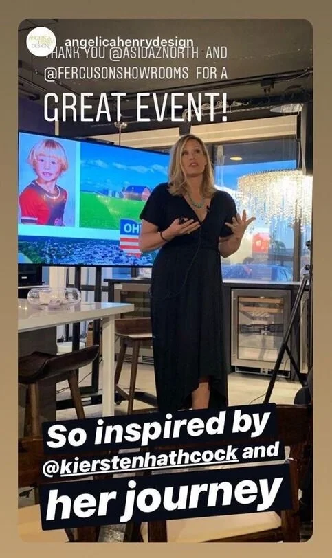 A woman standing in a restaurant or event space giving a speech or presentation. Behind her is a large screen displaying an animated child with blonde hair and a red shirt, and outdoor scenery. The space has large windows, a chandelier, and tables wi