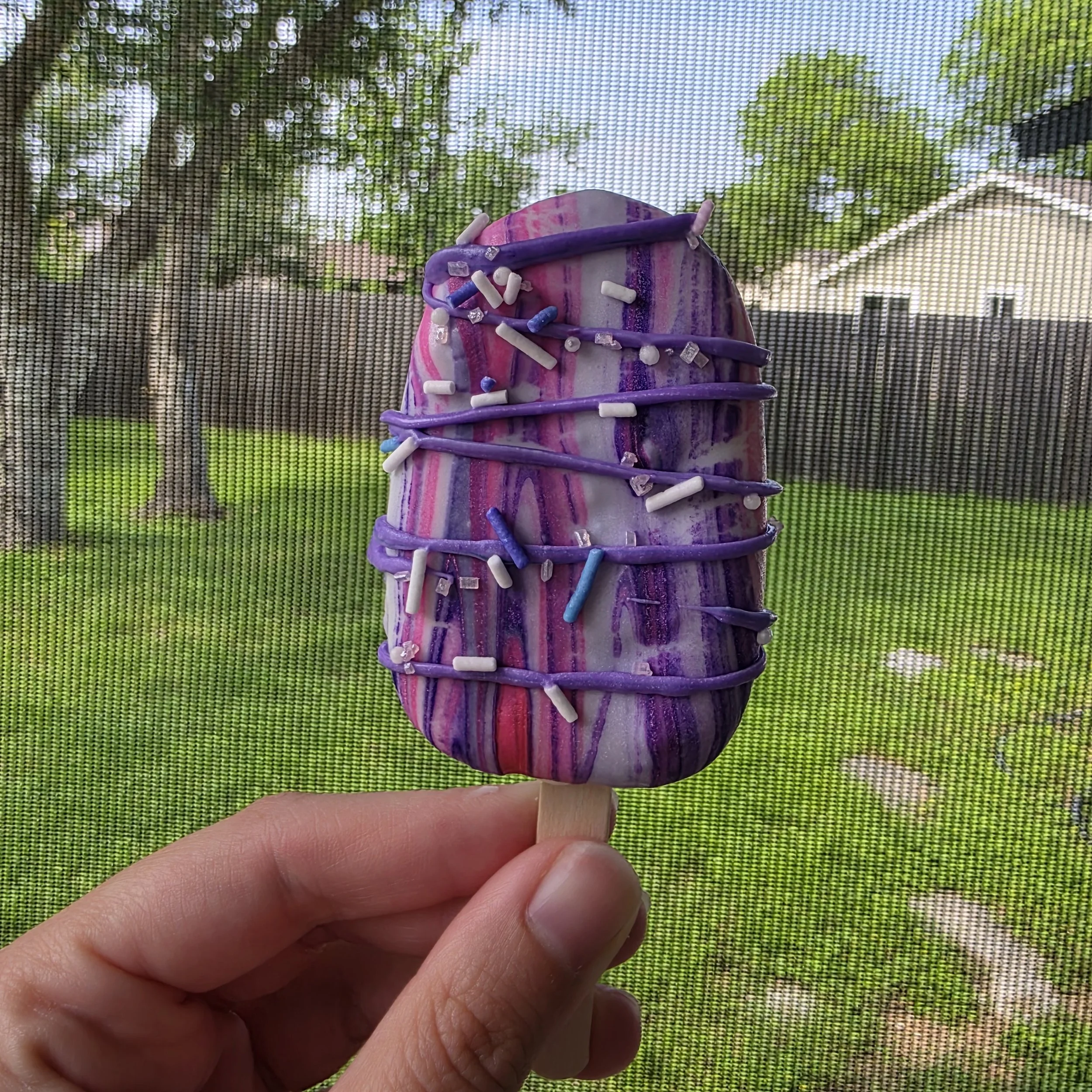 Close-up of a hand holding a colorful marble with purple, pink, and white streaks, decorated with purple and white tiny sprinkles, against a background of a mesh screen with trees and a house visible outside.
