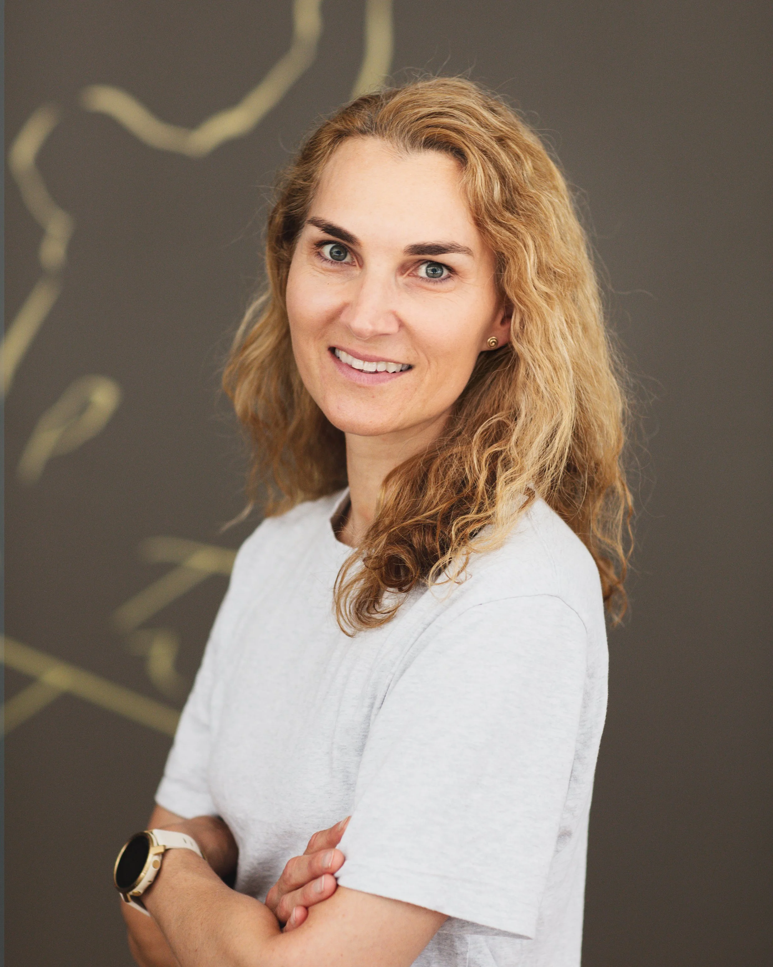 Portrait de Michelle Jaquet-Fertek, physiothérapeute et fondatrice de PhysioAir, portant un t-shirt blanc, portant une montre, souriante, contre un fond gris avec des motifs flous en beige. La femme regarde l'appareil photo.