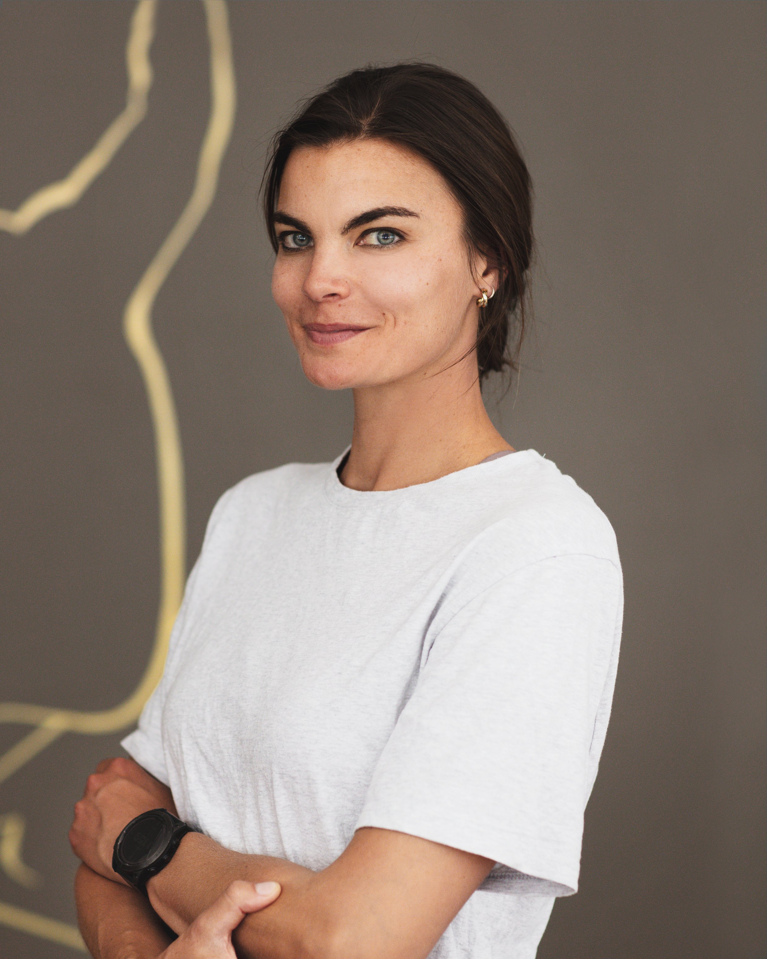 Portait de Justine Saint-Paul , physiothérapeute chez PhysioAir, souriant légèrement, portant un t-shirt blanc et un bracelet noir, croisée les bras, contre un fond gris.