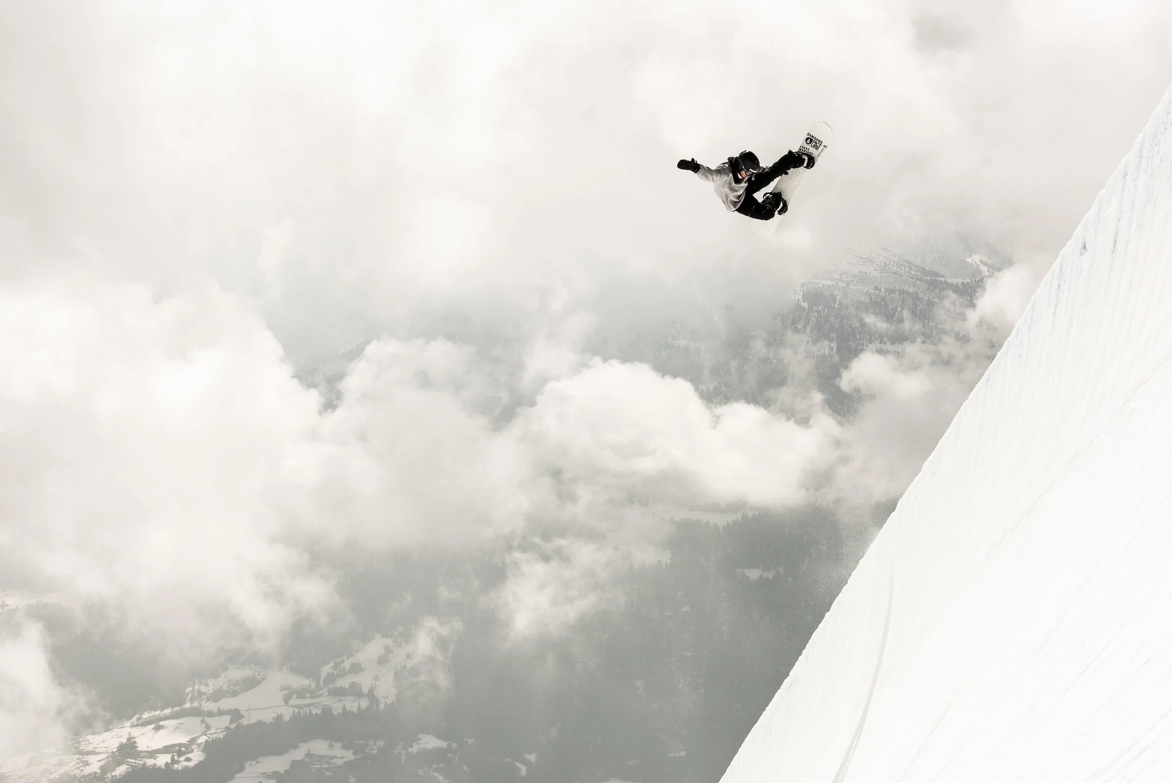Un snowboardeur effectue un saut dans un paysage enneigé avec des montagnes brumeuses en arrière-plan.