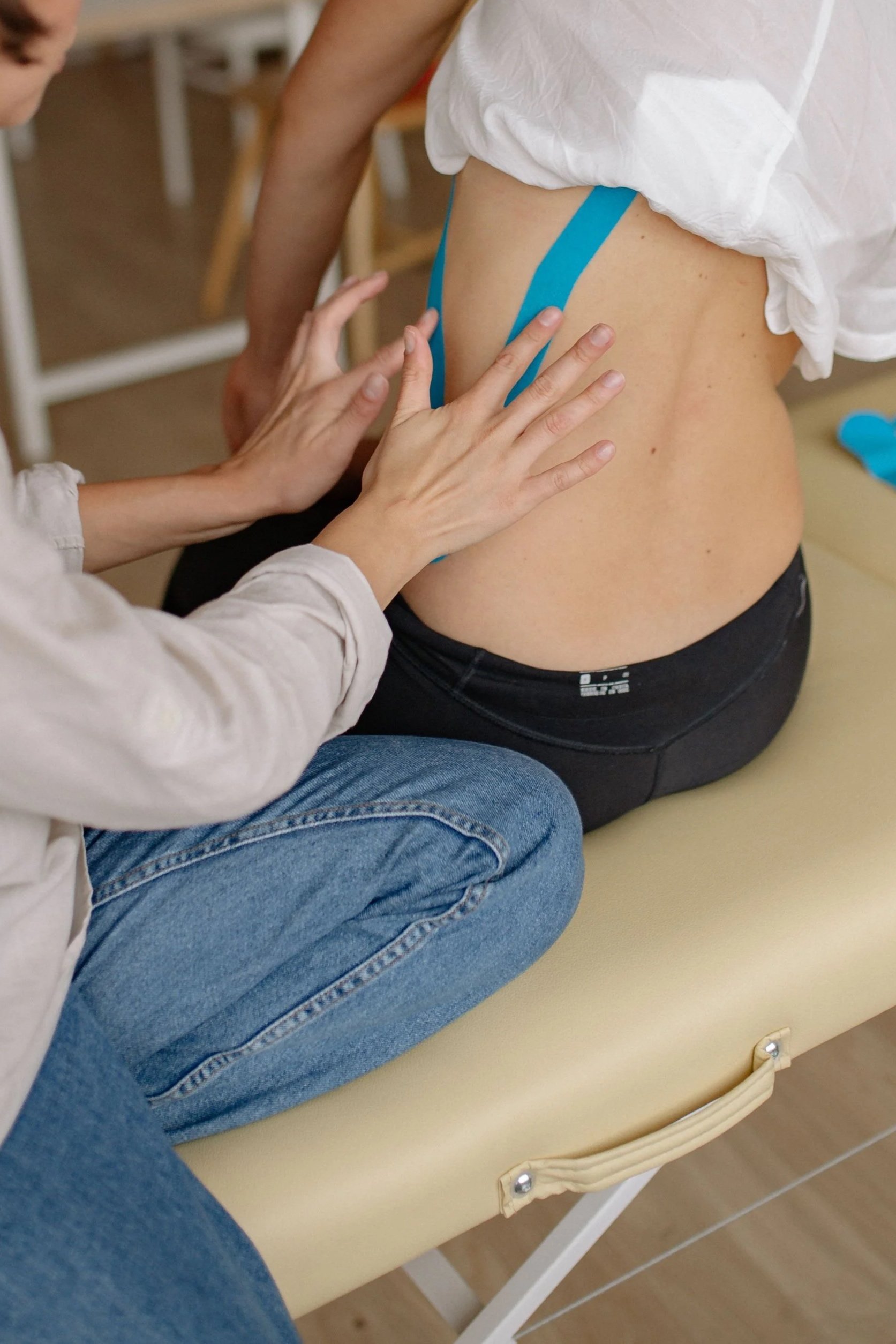 Une personne en train de faire un massage ou une thérapie sur le côté d'une autre personne qui porte un pantalon noir et un haut blanc, assise sur un fauteuil beige en milieu intérieur.