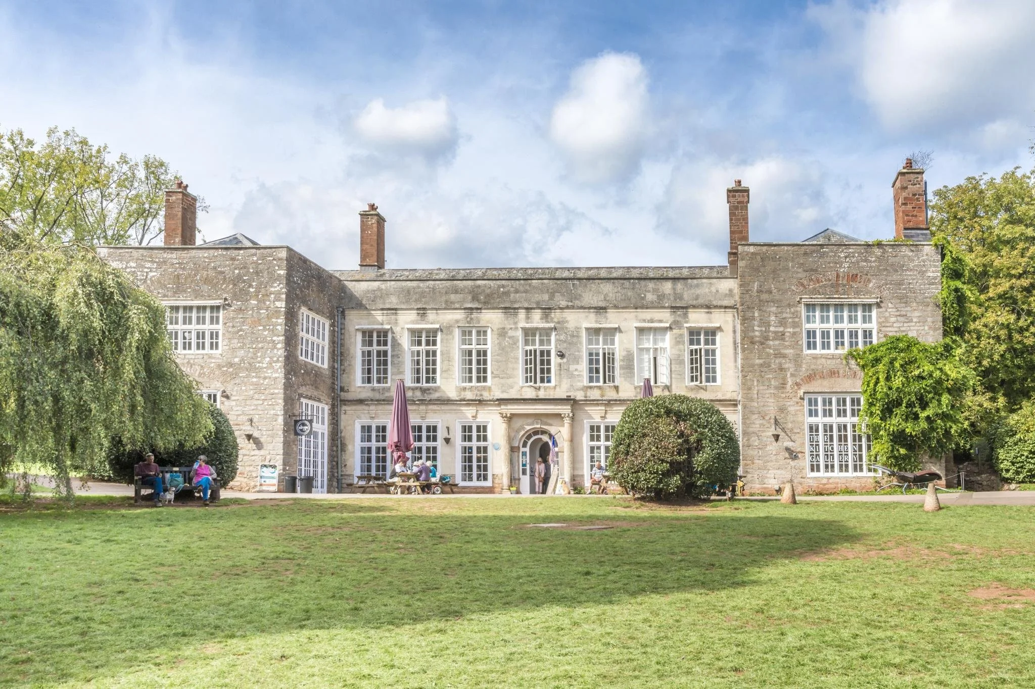 Historic stone building with multiple windows and three prominent chimneys, surrounded by greenery and trees, with people sitting on benches and walking in front.