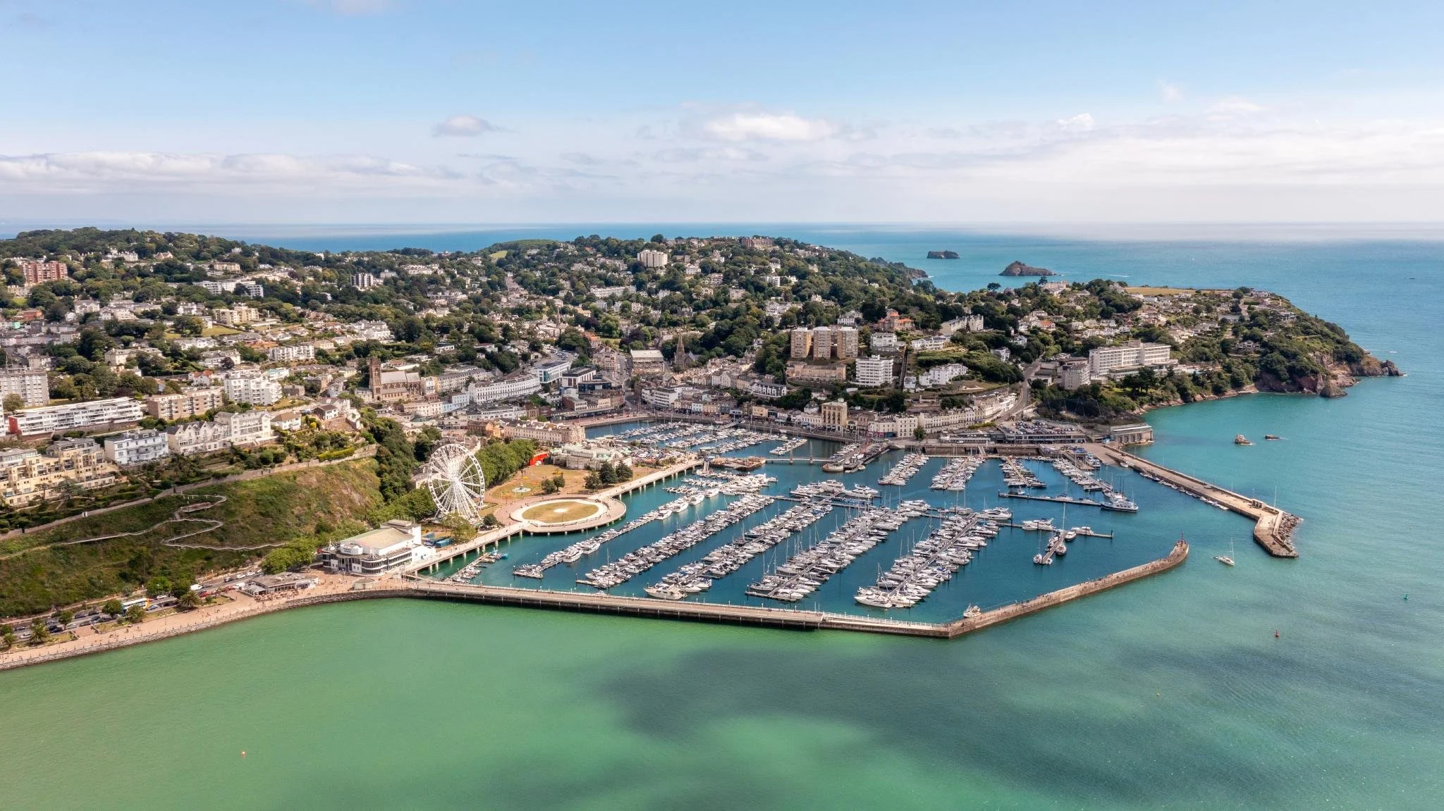 Aerial view of a coastal city with a harbor filled with boats, surrounding hills with residential and commercial buildings, a Ferris wheel, and the ocean in the background.