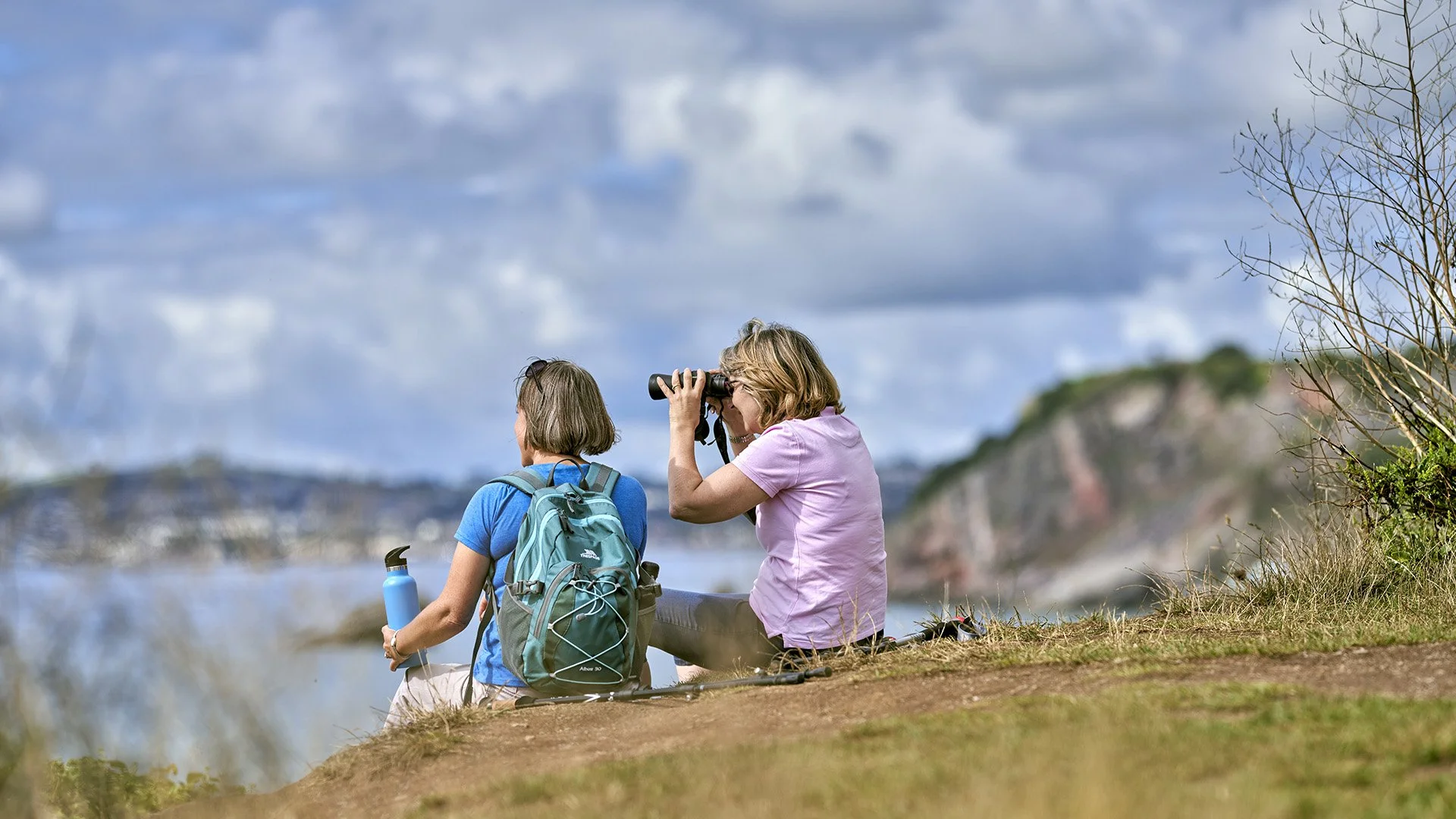 Two women sitting on a hillside near water, one looking through binoculars, the other holding a water bottle, with a backpack; in the background, hills, water, and cloudy sky.