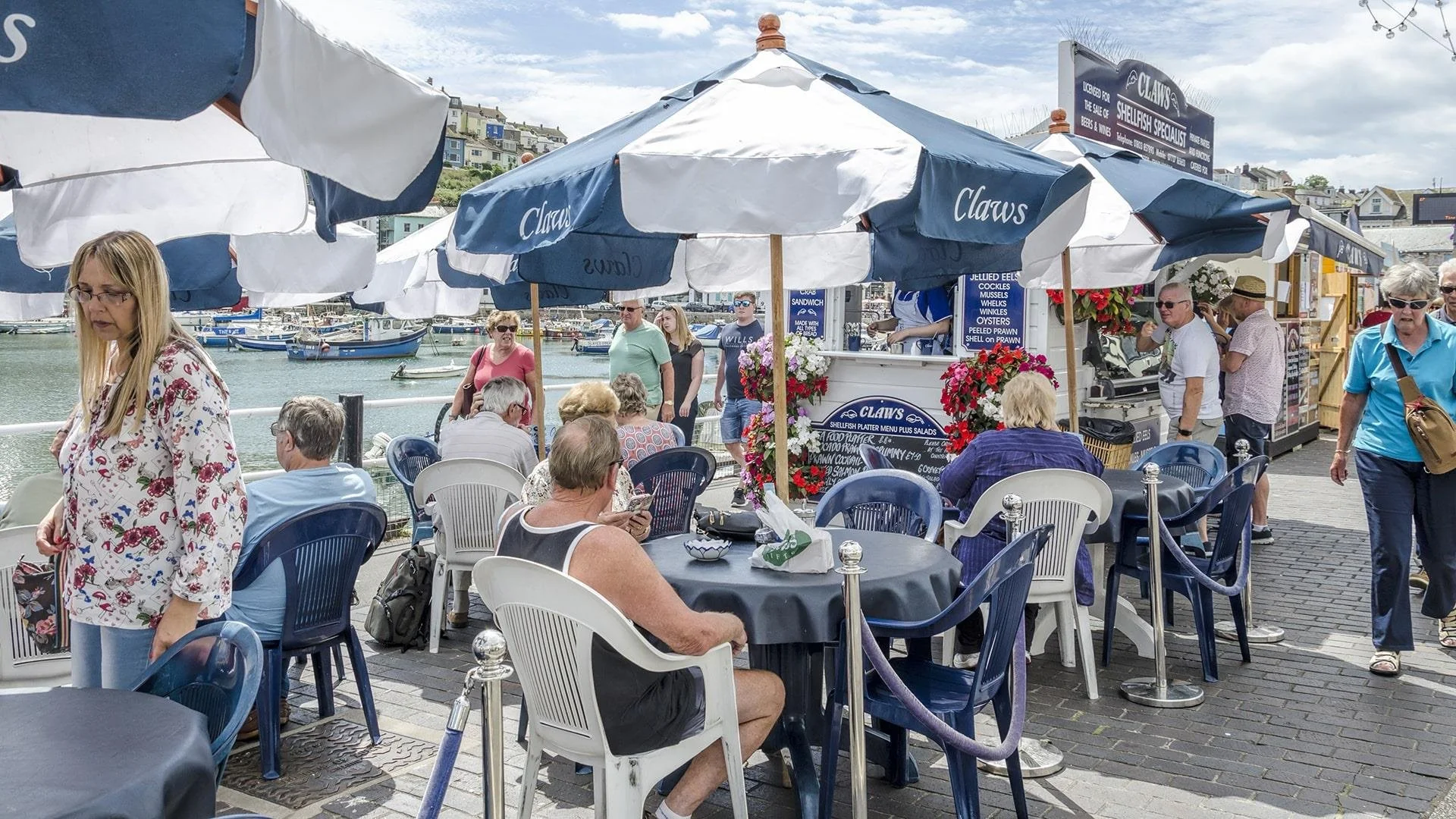 People dining outdoors at tables with blue and white chairs near a waterfront, with boats in the harbor, under umbrellas, some standing and some seated, with buildings and a cloudy sky in the background.