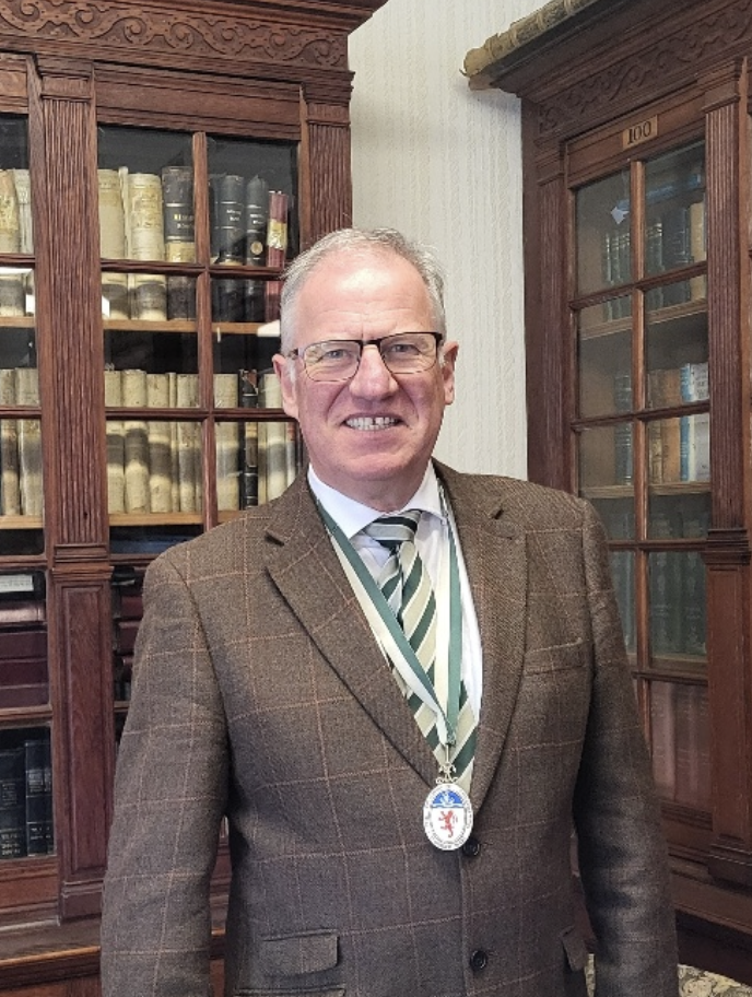A man wearing glasses and a brown plaid suit with a striped tie and a medal around his neck, standing in front of a wooden bookcase filled with books.