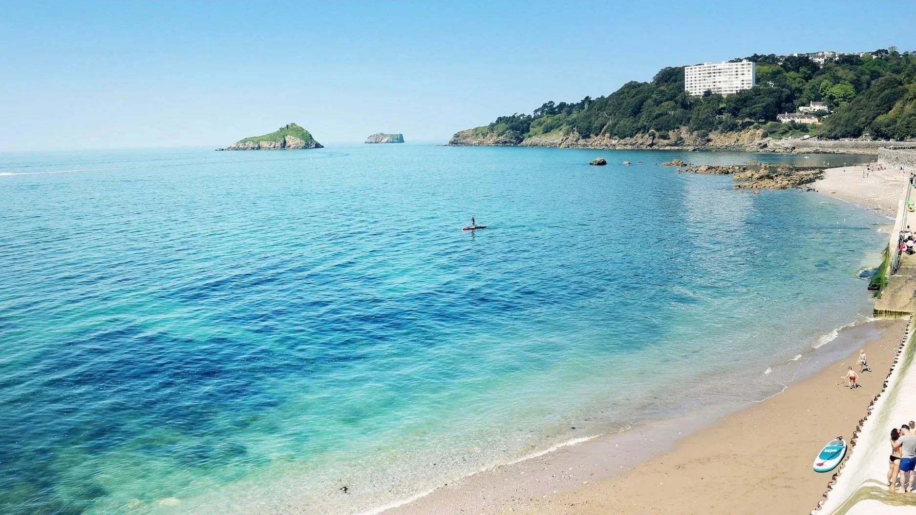 A view of a beach with clear blue water and sandy shore. There are a few people on the beach and a paddleboarder in the water. In the background, there are green hills with a white building on top.