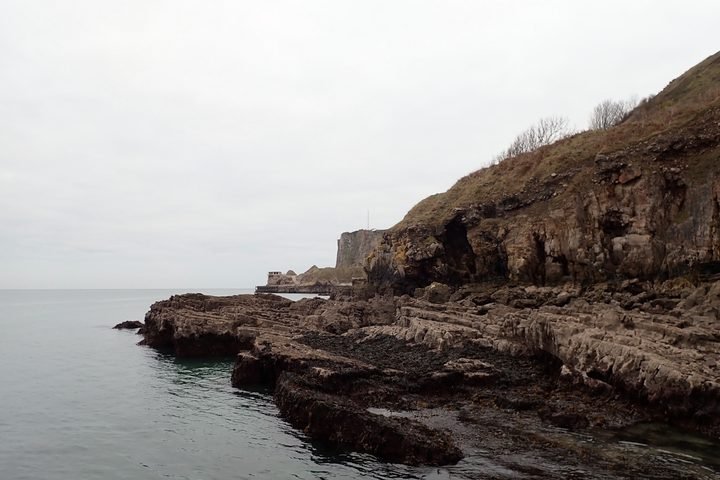 Overcast sky view of rocky coastal cliffs with layered rock formations and a small building at the far end of the coastline.