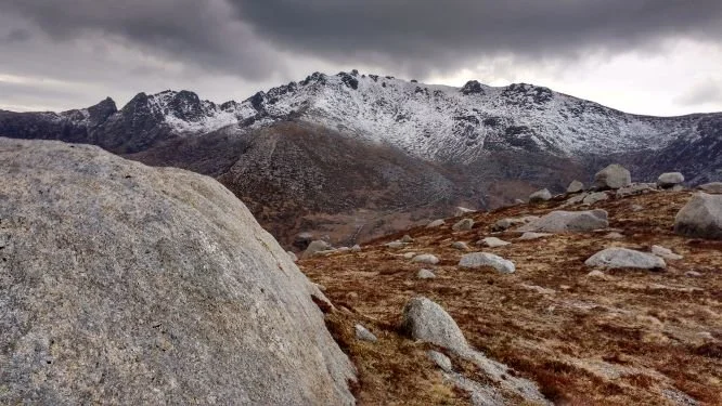 Mountains with snow-capped peaks and rocky terrain under dark, cloudy sky.