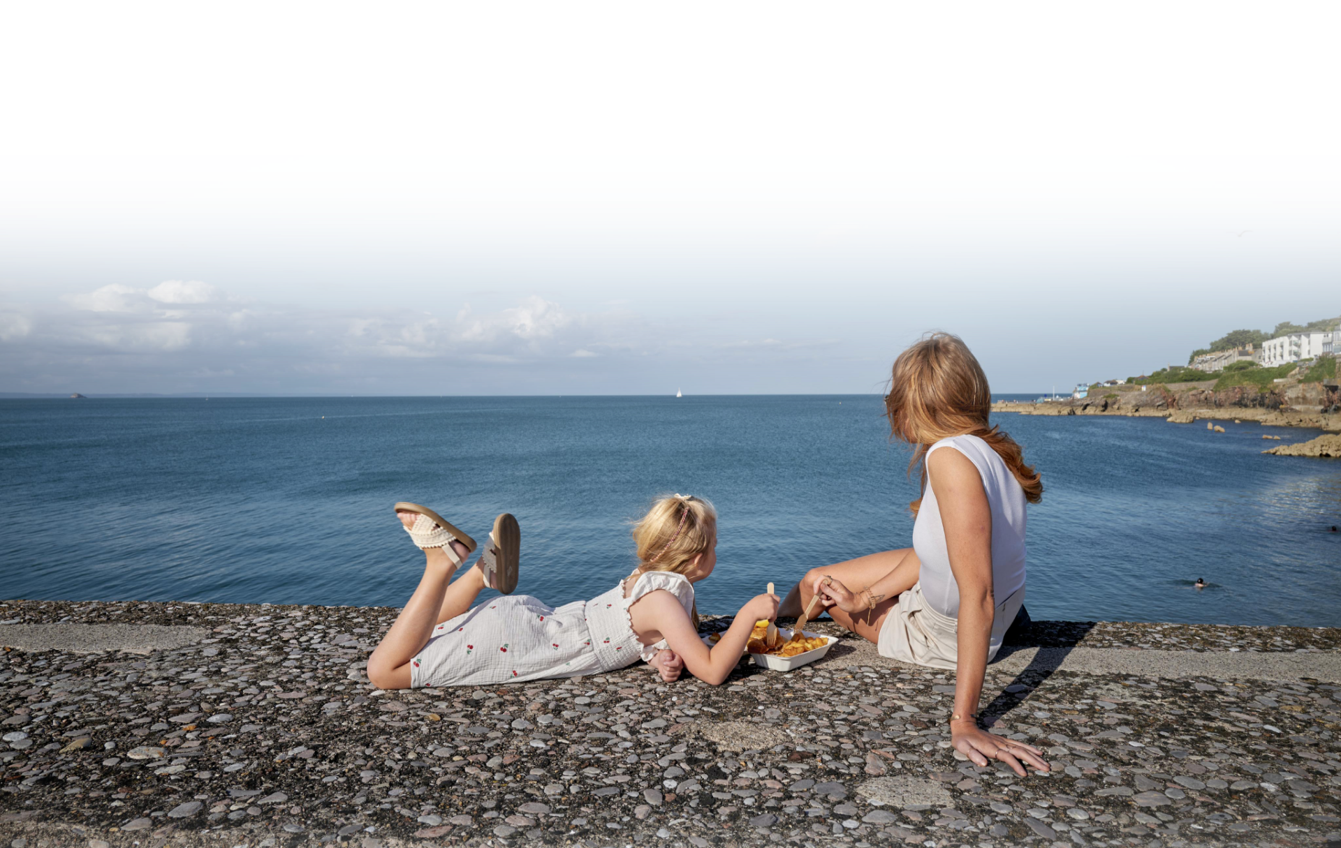 A woman and two young girls relaxing by the water on a rocky shore. The woman is sitting and holding the hand of one girl, while the second girl is lying on her stomach, wearing sandals. The scene features an ocean with sailboats, a cloudy sky, and coastal buildings in the background.